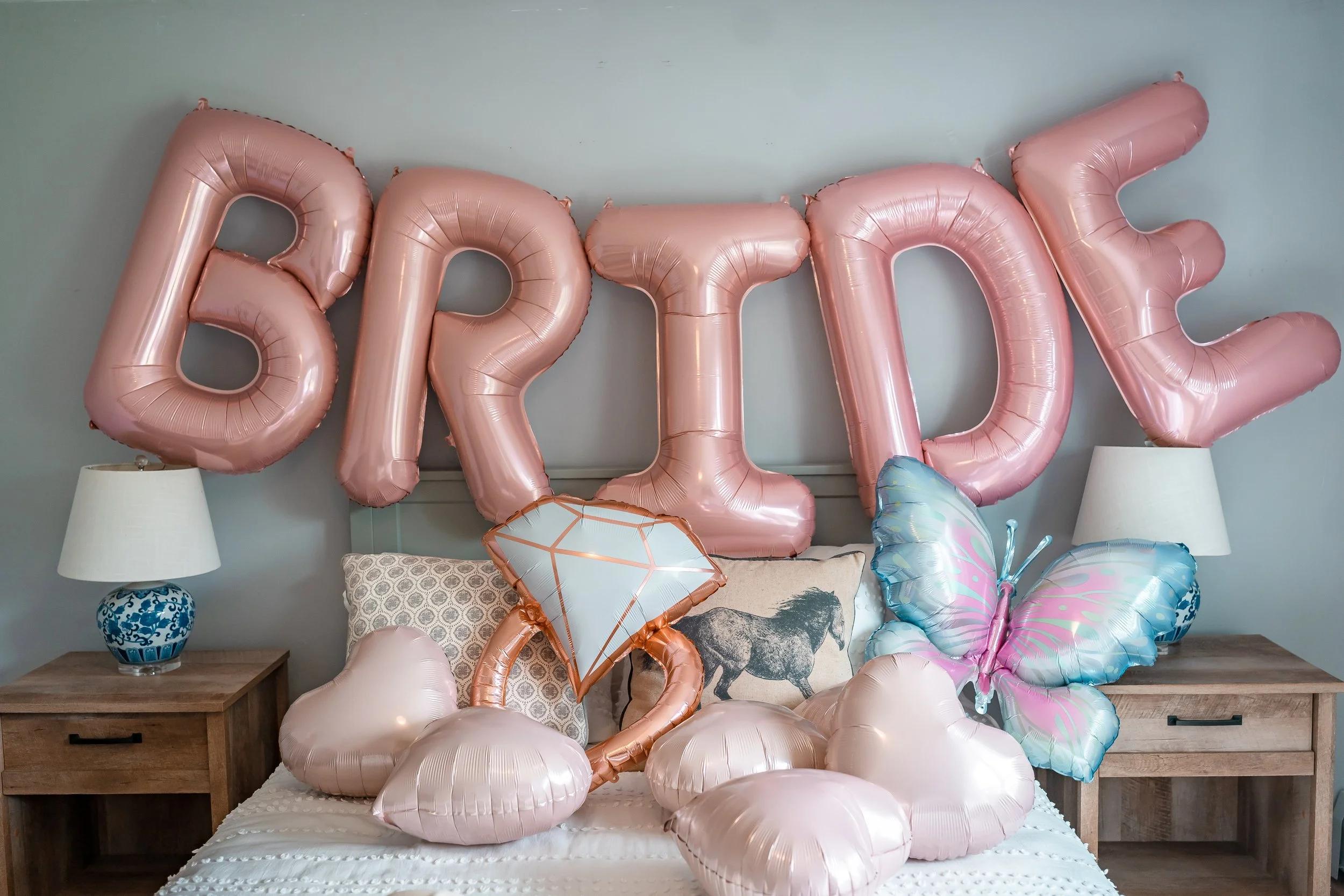 Pink balloon letters spelling 'BFF' on a bed with heart-shaped and diamond-shaped balloons, a pastel butterfly balloon, and pillows, flanked by bedside lamps.