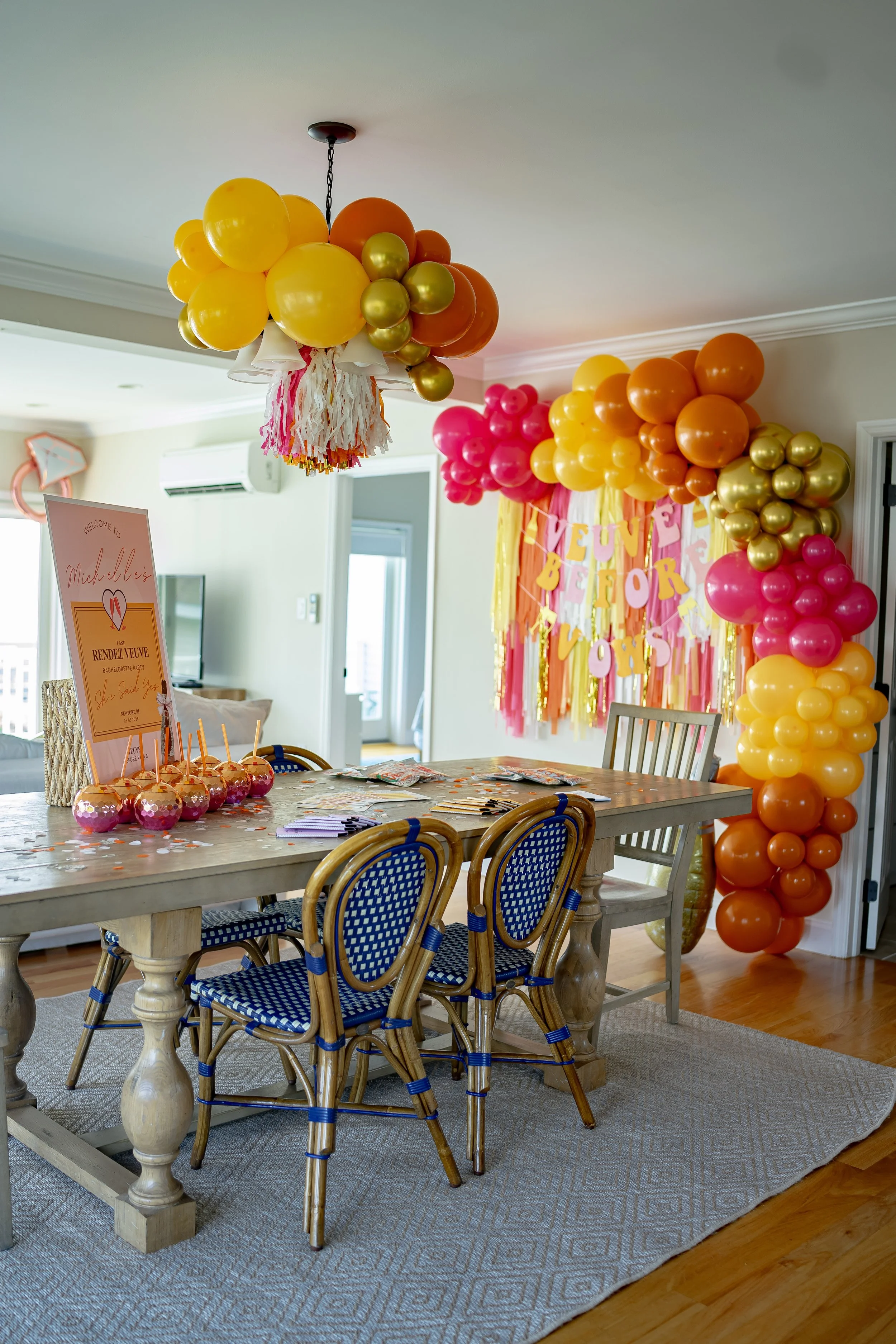 Party room decorated with yellow, orange, pink, and gold balloons, a colorful backdrop with streamers and a banner, and a wooden table with party favors and a sign.