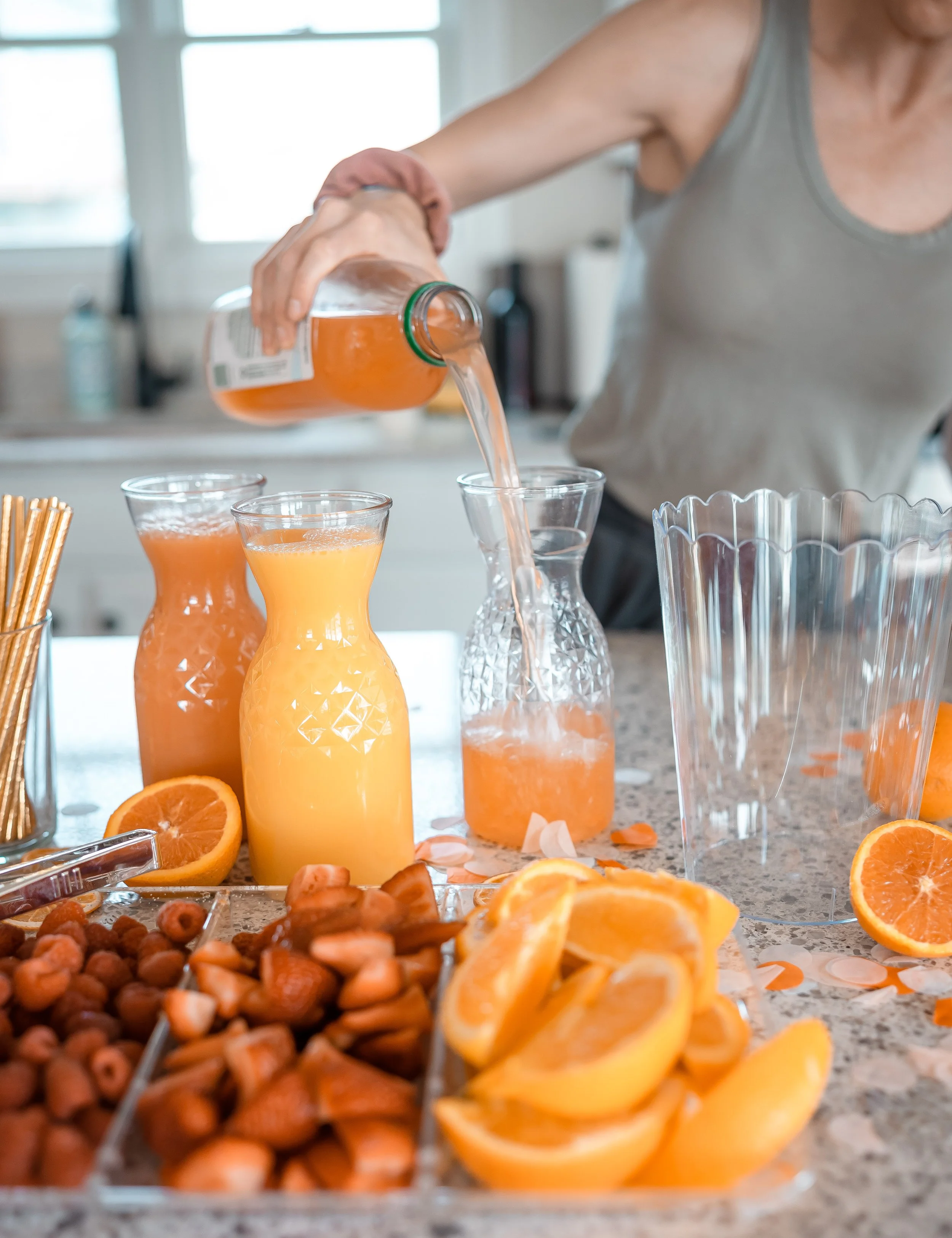 Person pouring orange juice into a glass on a kitchen counter with bottles of juice, orange slices, and fruit snacks.