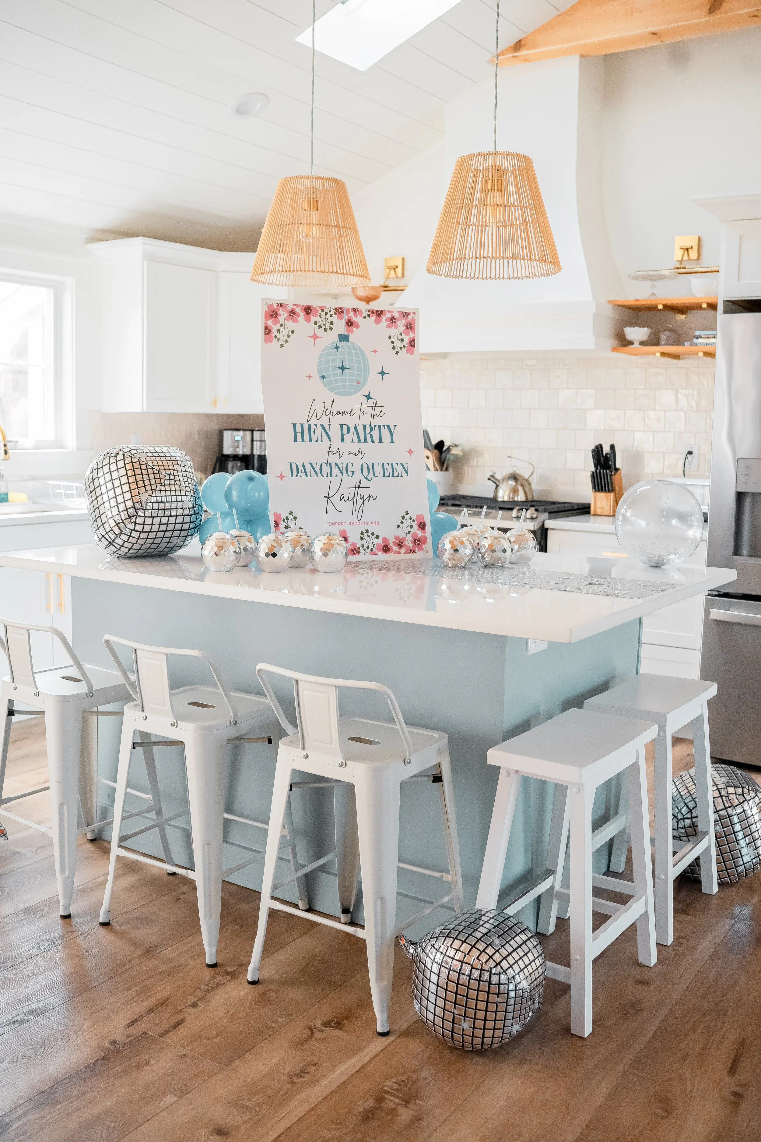 A decorated kitchen island with a sign that reads 'Welcome to the Hen Party, for our Dancing Queen, Kaitlyn.' The island has teal painted sides, and is surrounded by white stools and a small white step stool. Balloons and metallic disco ball decor ar