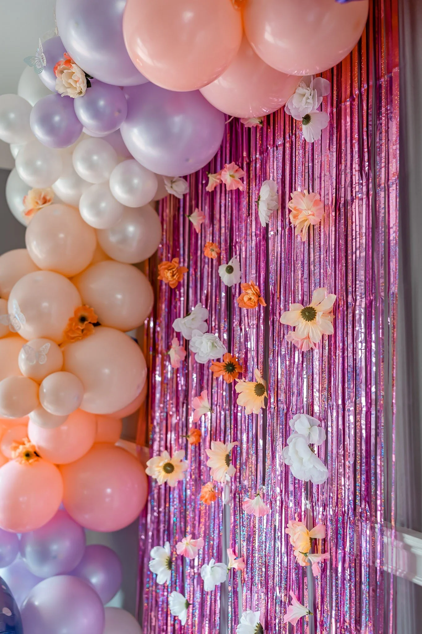 Decorative setup featuring pink, purple, and white balloons arranged on the left side and a pink foil fringe curtain with hanging paper flowers and butterflies on the right side for a celebration or party.