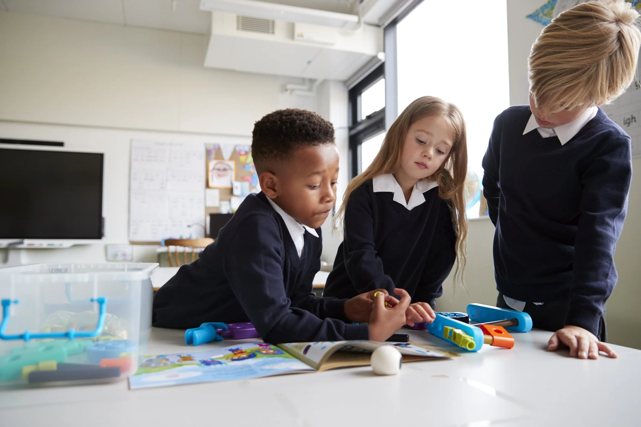 Three children in school uniforms working together at a table with educational toys and a magazine in a classroom.