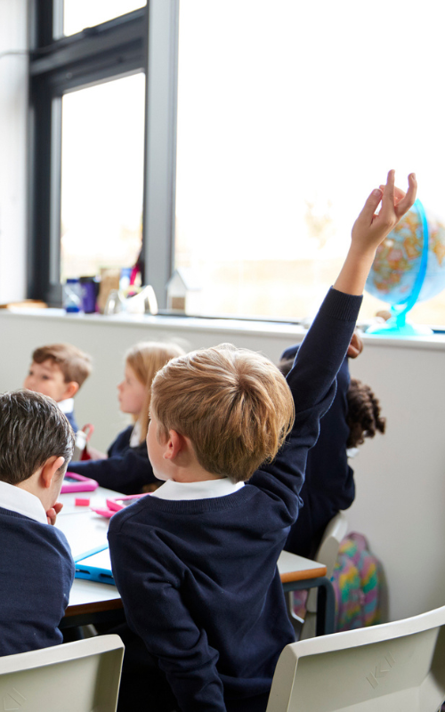 A young boy in a classroom raising his hand while sitting at a desk with other students, with a window and a globe in the background.