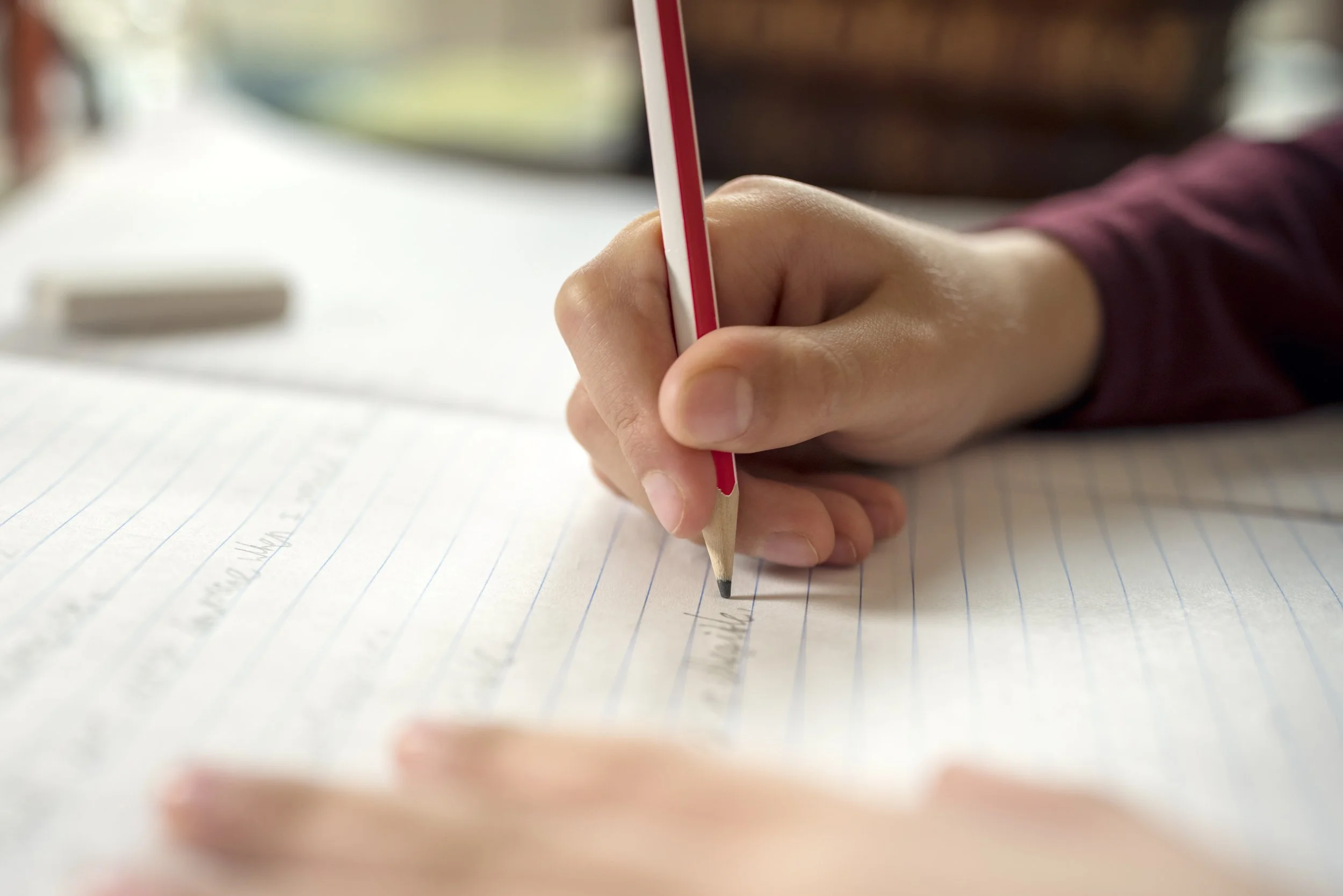 Child writing on a white sheet of paper with a blue pen at a desk with notebooks, a yellow tape measure, and an eraser.