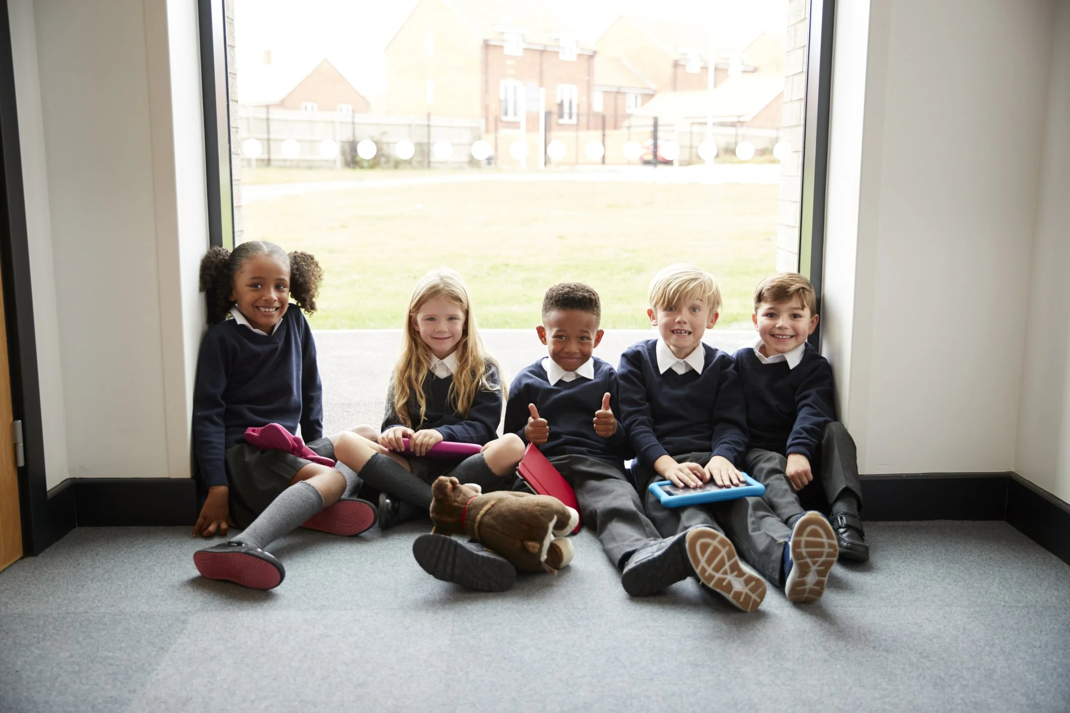 Group of five school children sitting on the floor next to a door, outside view of a grassy area and houses in the background