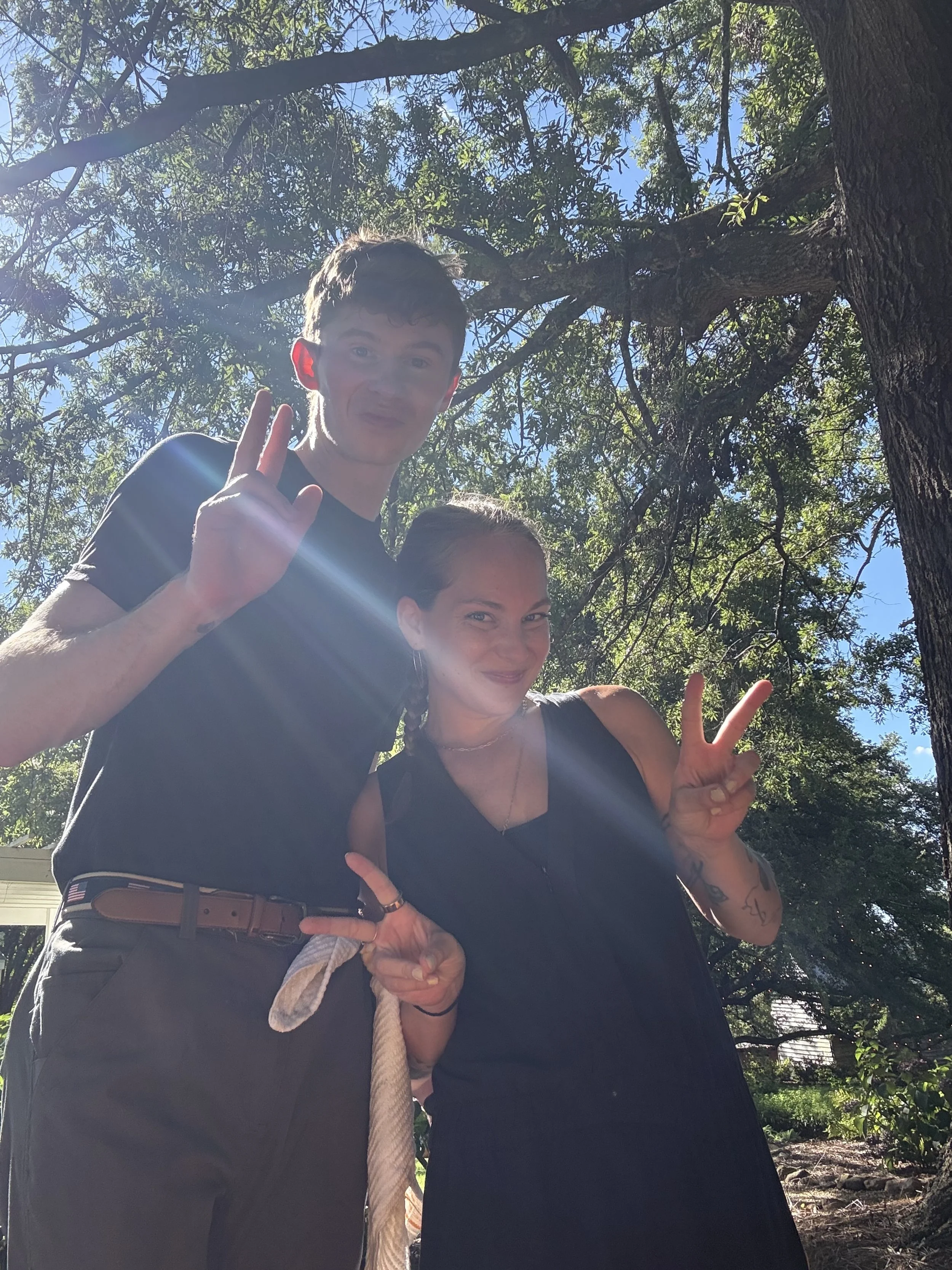 A young man and woman posing outdoors under a large tree, both smiling and making peace signs with their hands. The sun shines through the branches, creating a lens flare in the image.