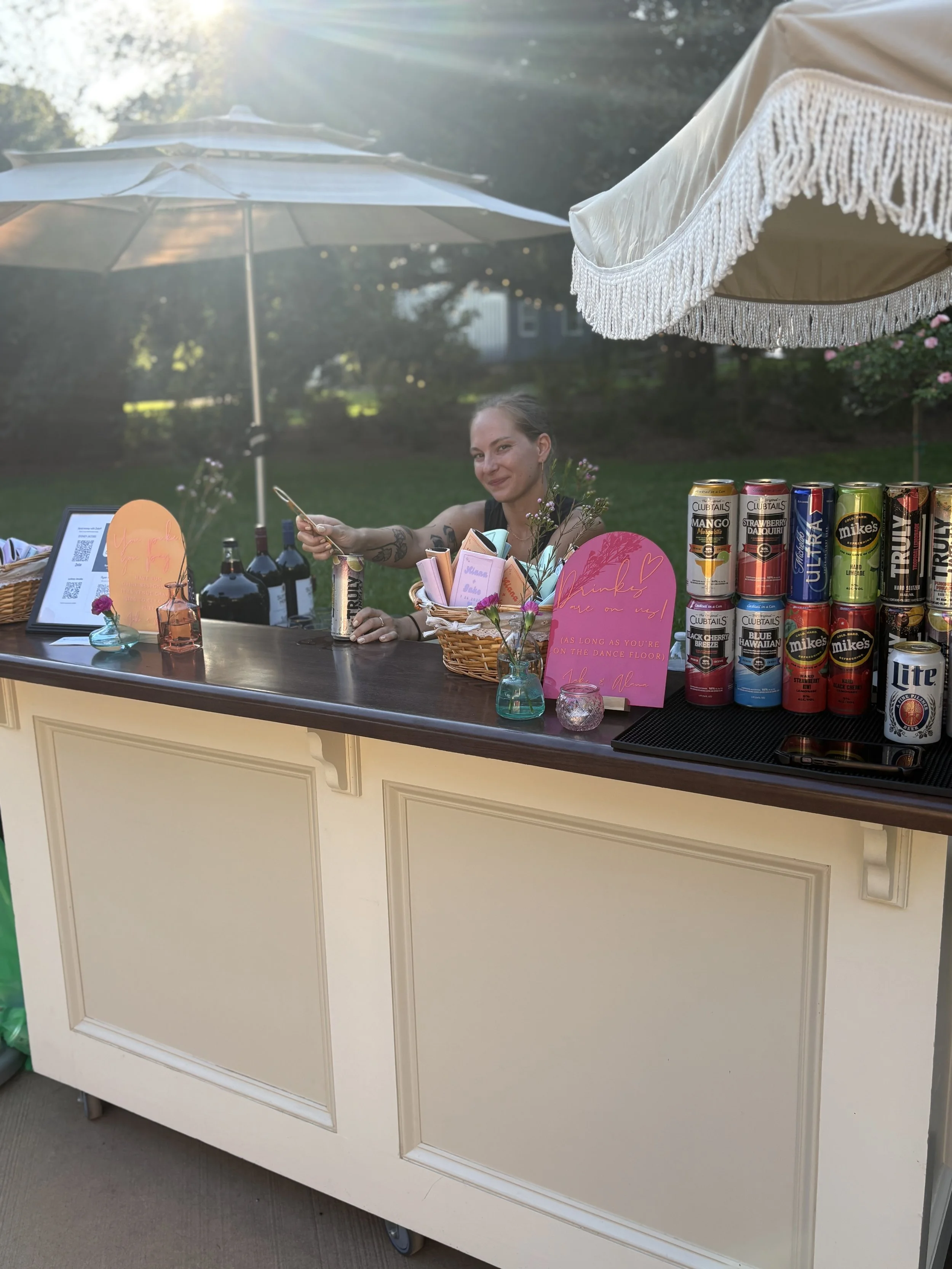A woman serving drinks at an outdoor bar during sunset, with wine bottles, canned beverages, and pink notes on the counter.