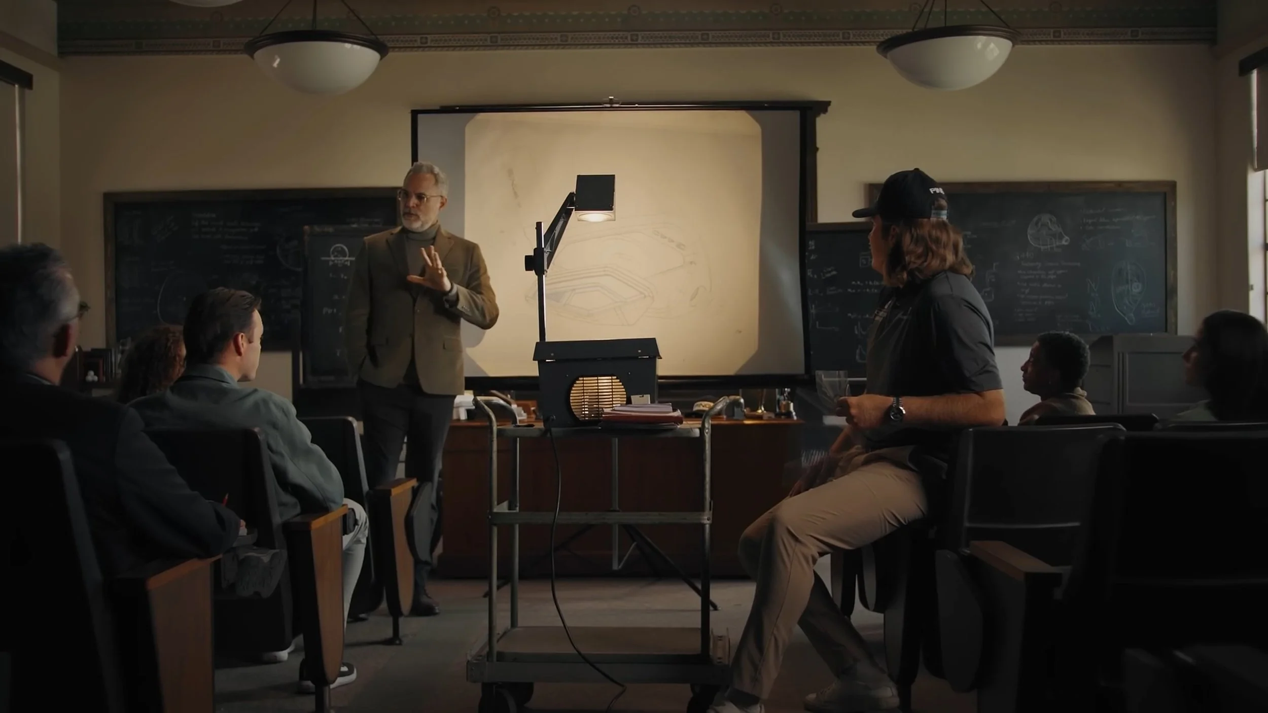 From Chariot's Advertisement with PING, a golf club company. A man in a tan blazer and glasses is giving a presentation in a classroom with a projector screen behind him. Several students are seated. The room has blackboards with sketches and notes.