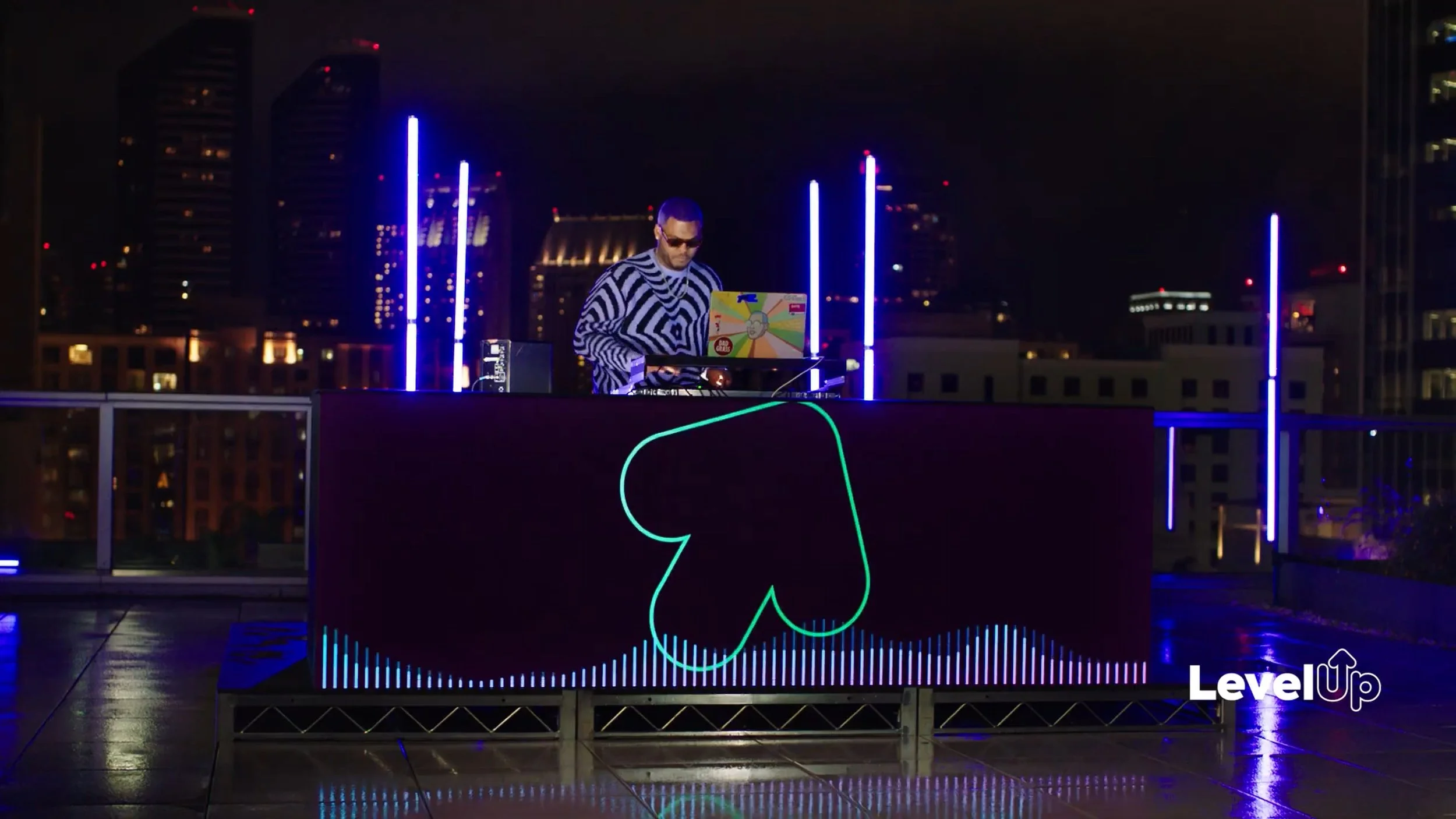 From Chariot's advertisement with ClickUp.A DJ wearing sunglasses is performing on a rooftop at night with city buildings in the background. Vertical blue neon lights decorate the scene, and the word 'LevelUp' is displayed in the bottom right corner.
