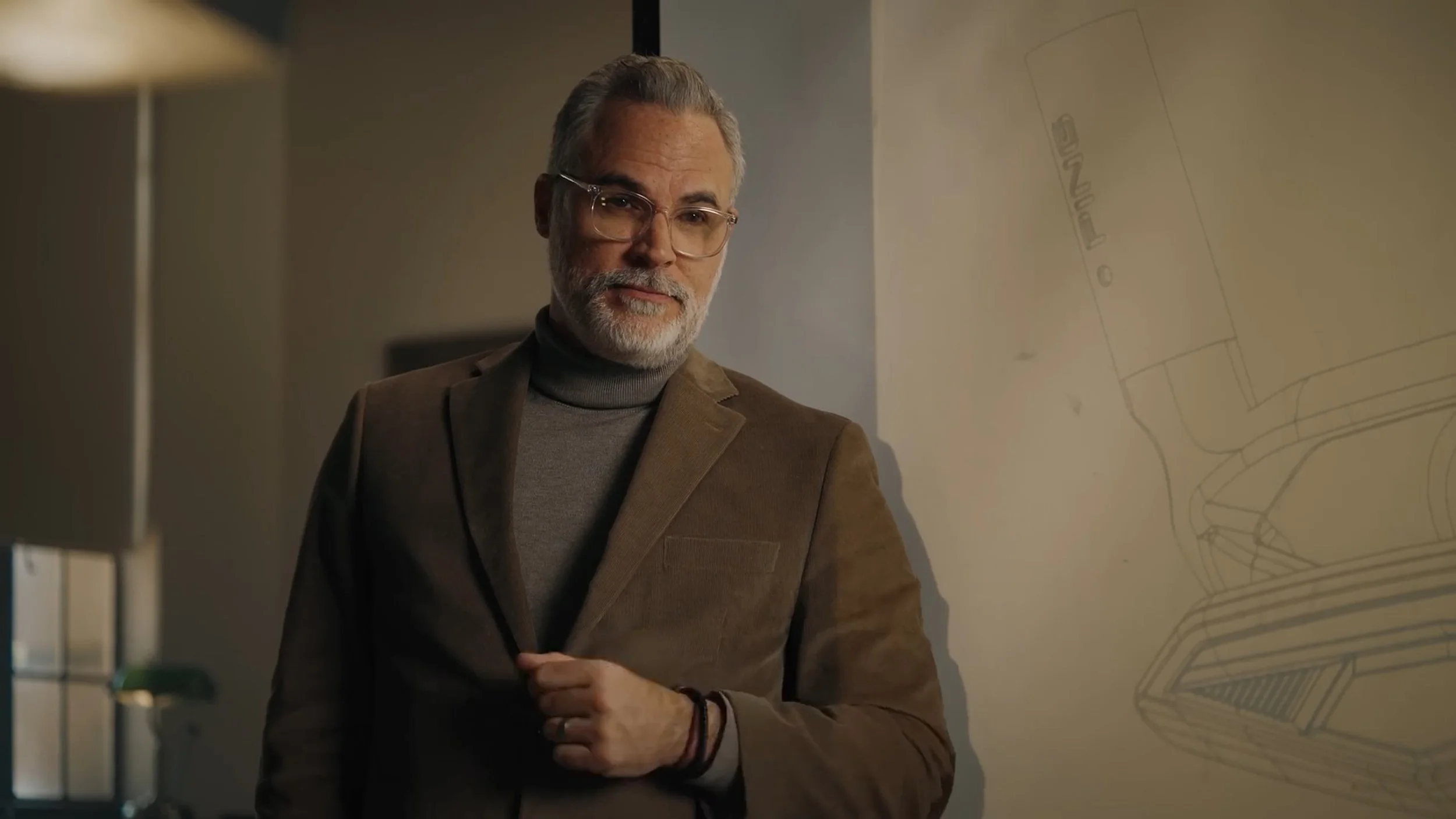 From Chariot's advertisement with PING, a golf club company. An older man with gray hair and beard wearing glasses, a brown blazer, and a gray turtleneck, standing indoors near a wall with a faint outline of a car drawing.