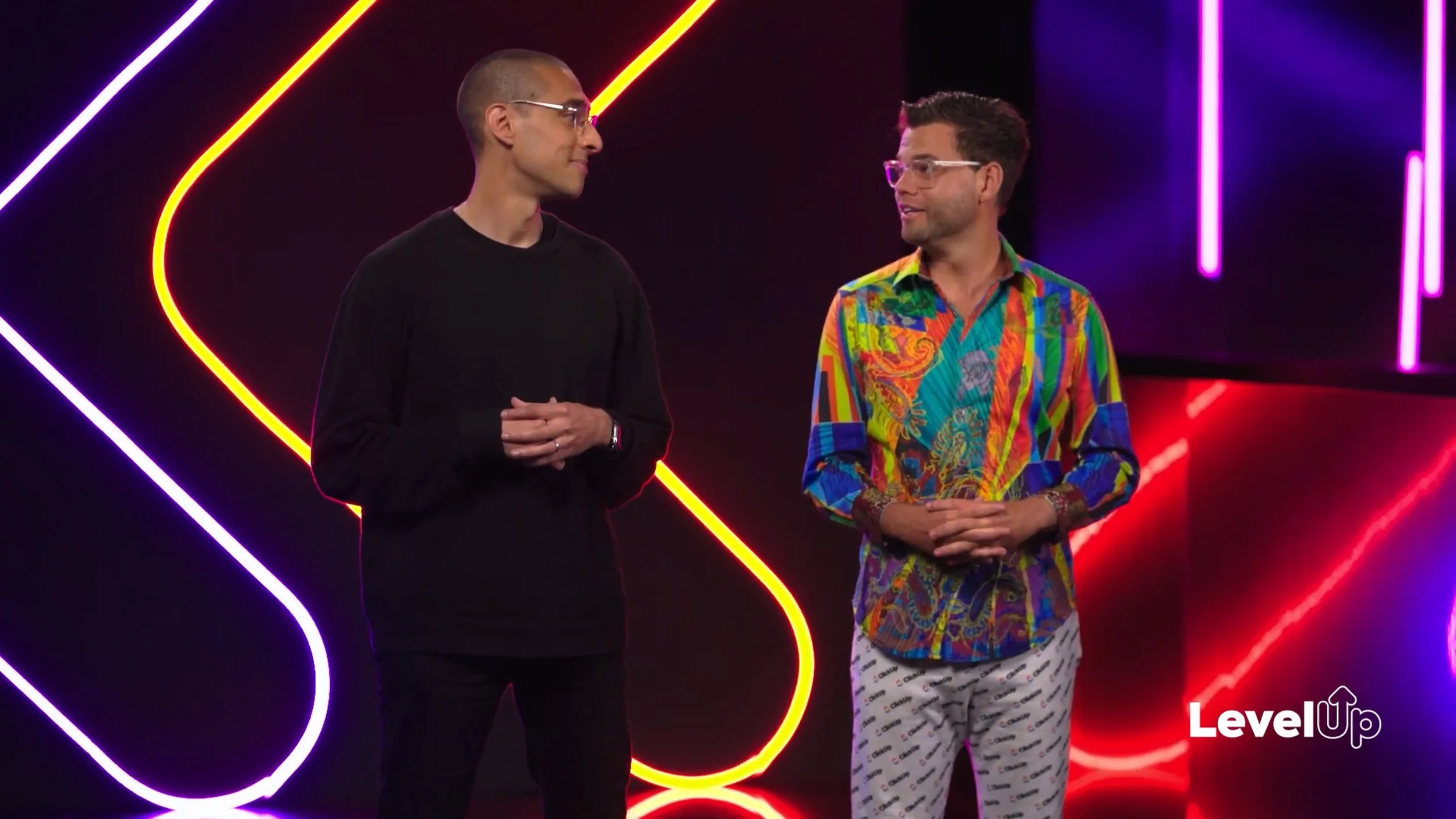 From Chariot's advertisement with ClickUp.  Two men standing on a stage with neon lights in the background, one wearing an all-black outfit and the other wearing a colorful patterned shirt and white pants with text.