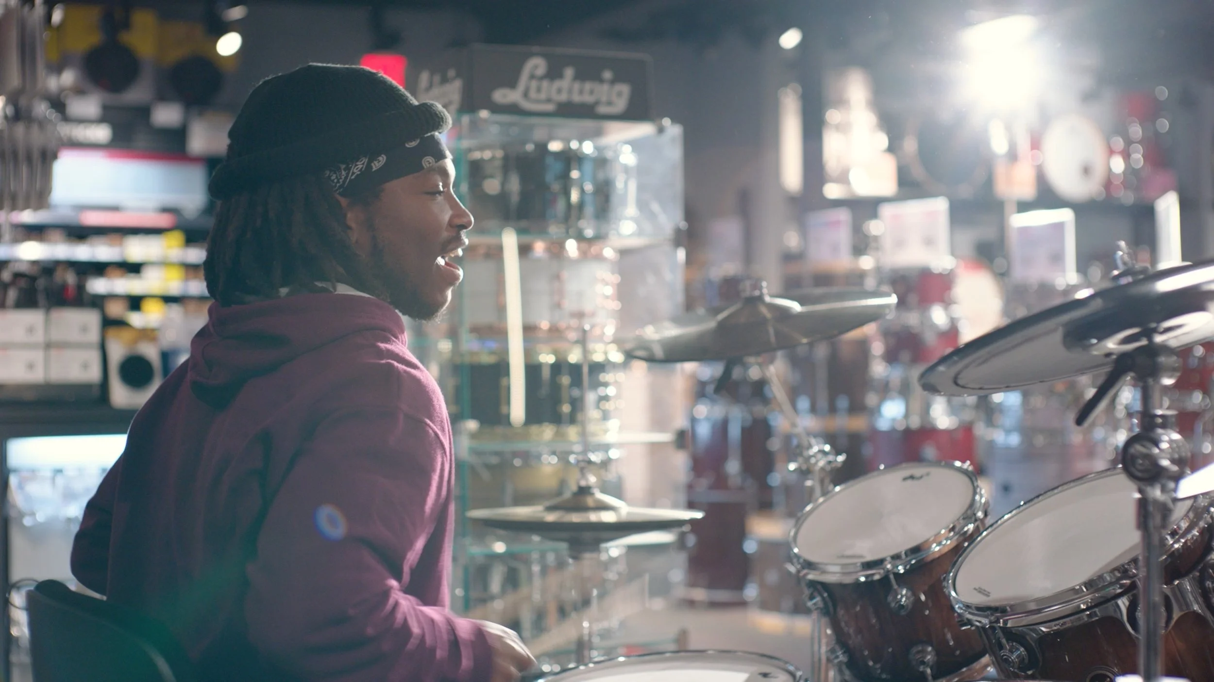 From Chariot's advertisement with Guitar Center. A man with dreadlocks wearing a black bandana and maroon hoodie playing a drum set inside a music store.