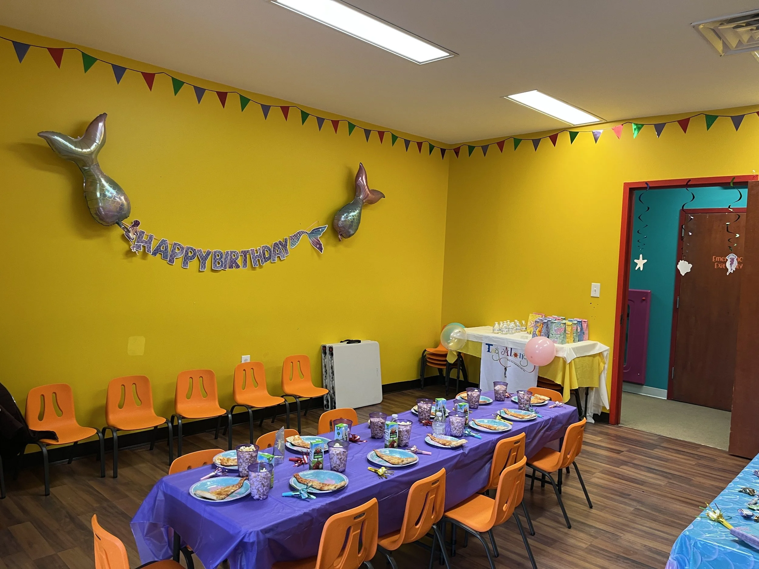 Colorful birthday party room with yellow walls decorated with balloons, a 'Happy Birthday' banner, and a table set with plates, cups, and party favors for children.