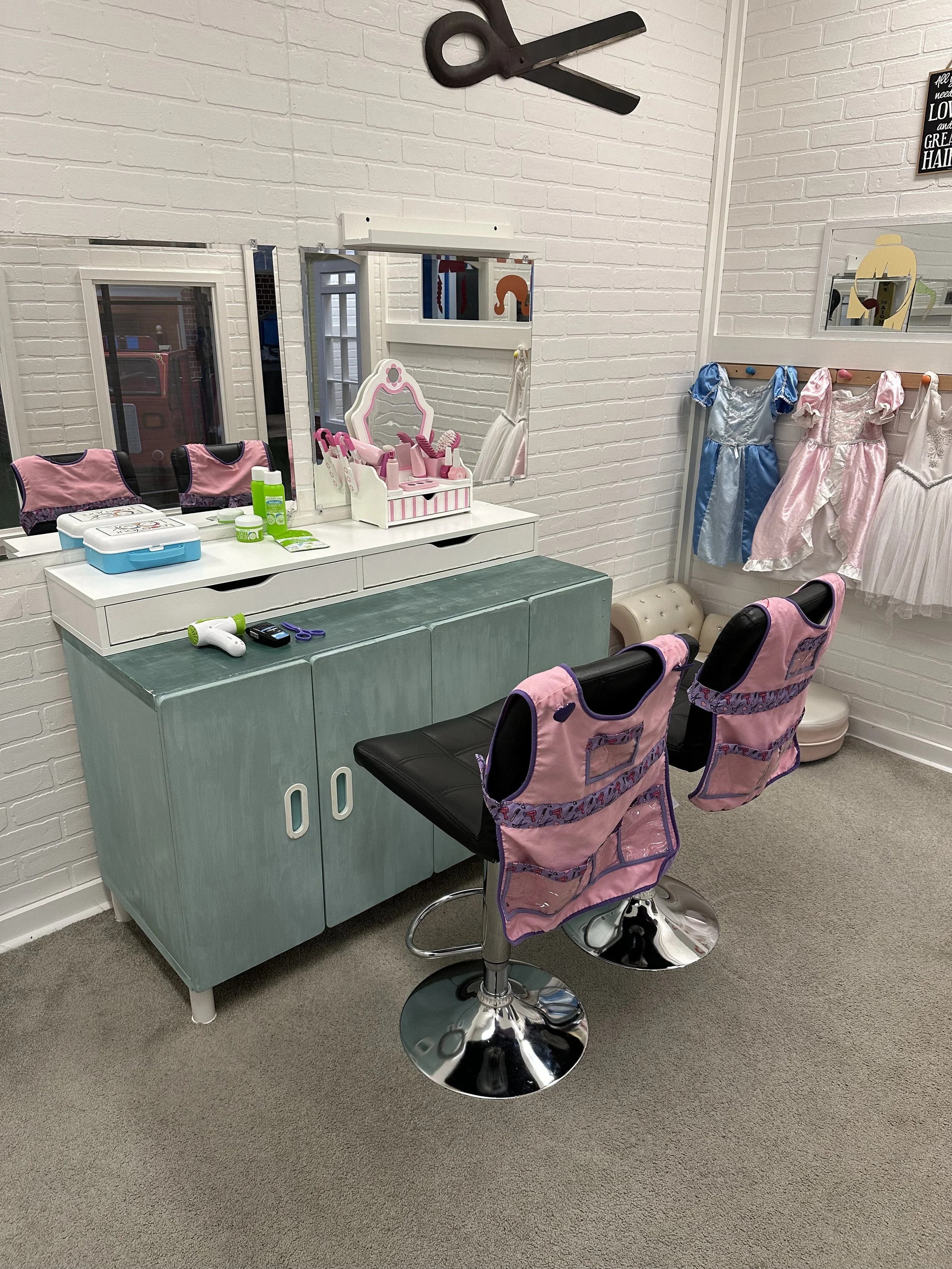 A children's dress-up and grooming area with two pink chairs, a dresser, and a mirror. There are children's dresses hanging on a rack, a small beige couch, and makeup items on the dresser. A wall decor with a pair of scissors and a mirror is also visible.