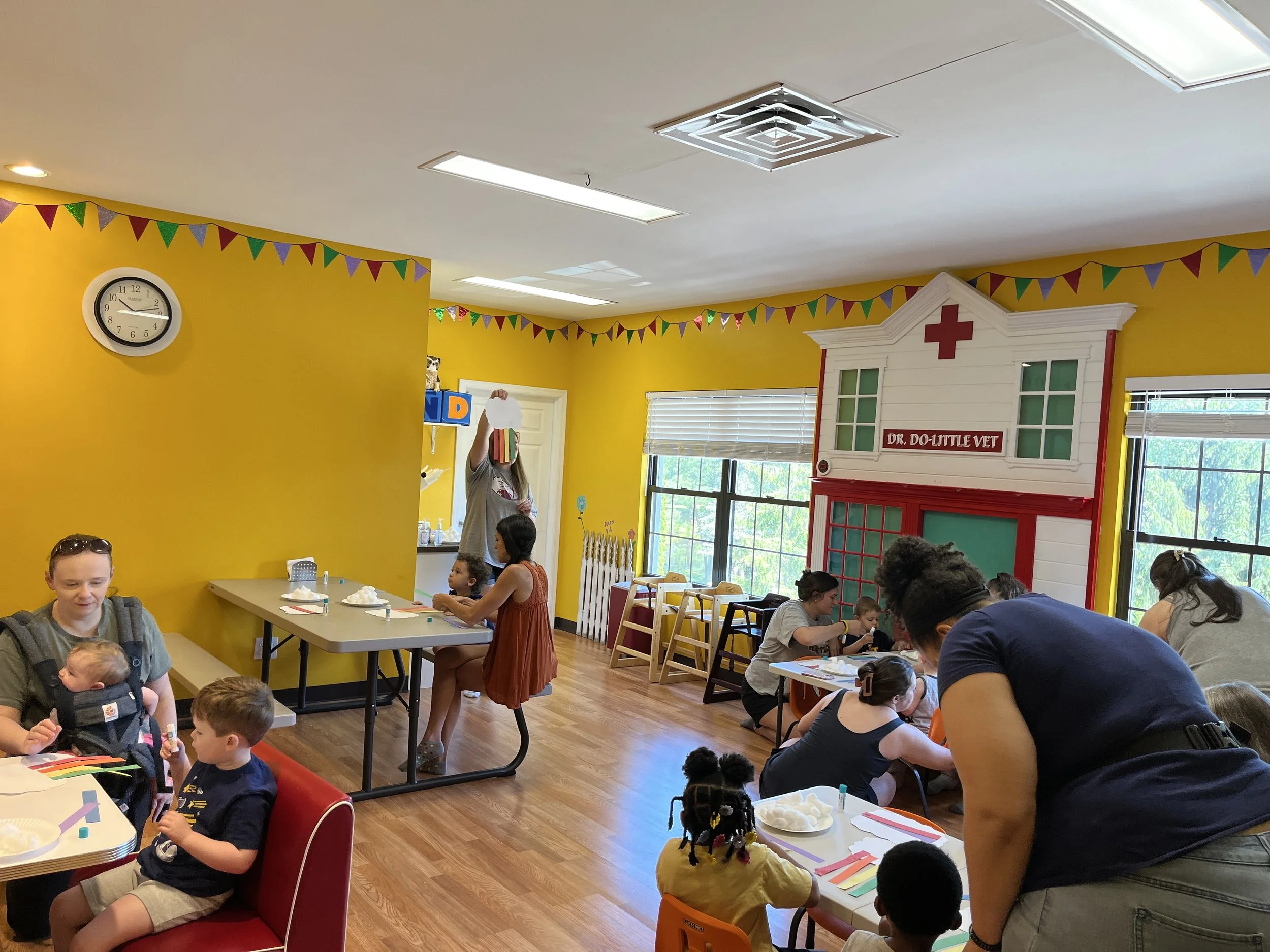 Children and adults in a colorful playroom decorated with bunting, and a large toy house labeled 'Dr. Do-Little Vet' in the background, engaging in activities around tables.
