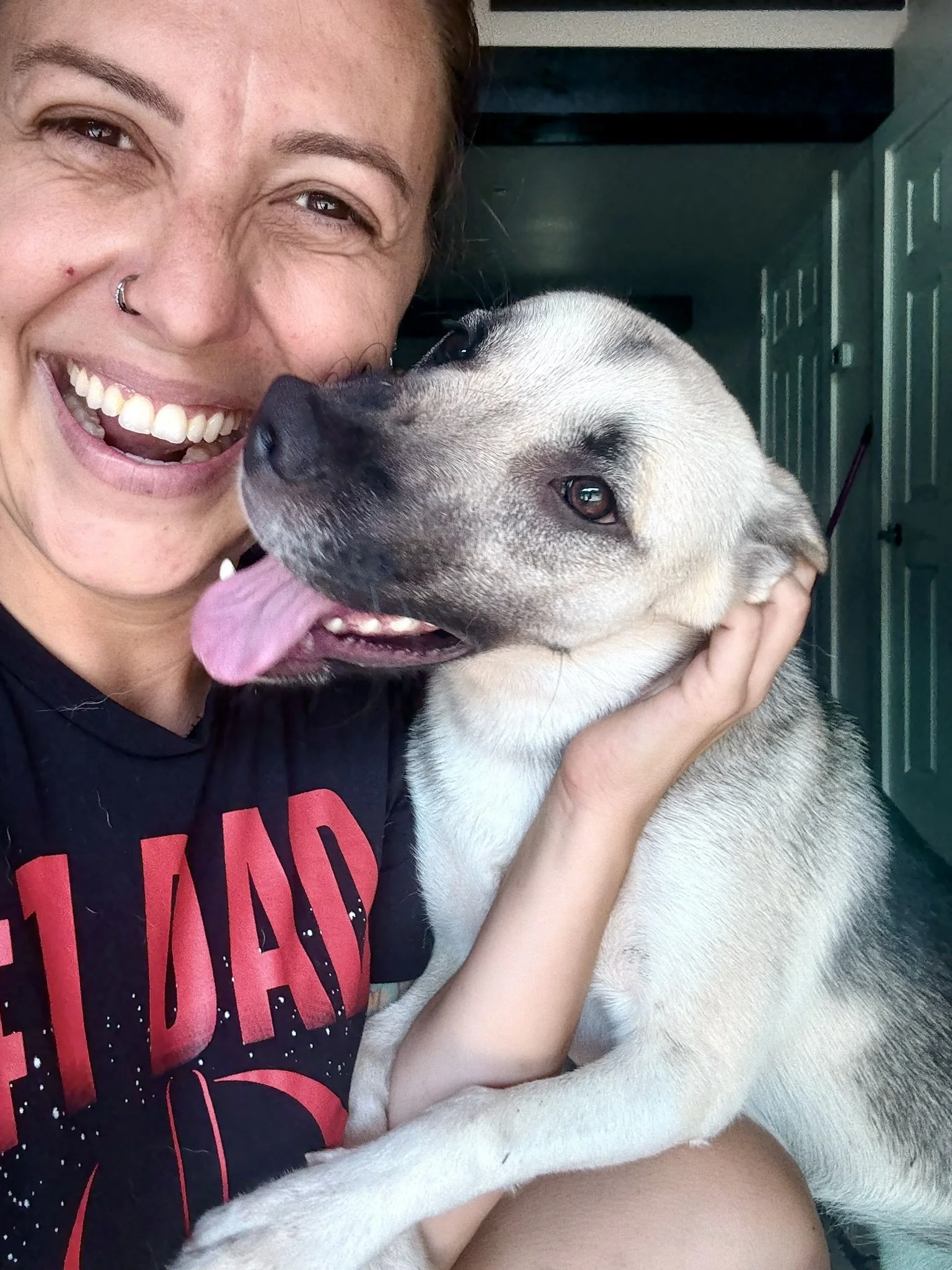 A woman with a nose ring smiling and holding a happy, light-colored dog with a dark snout and open mouth showing tongue.