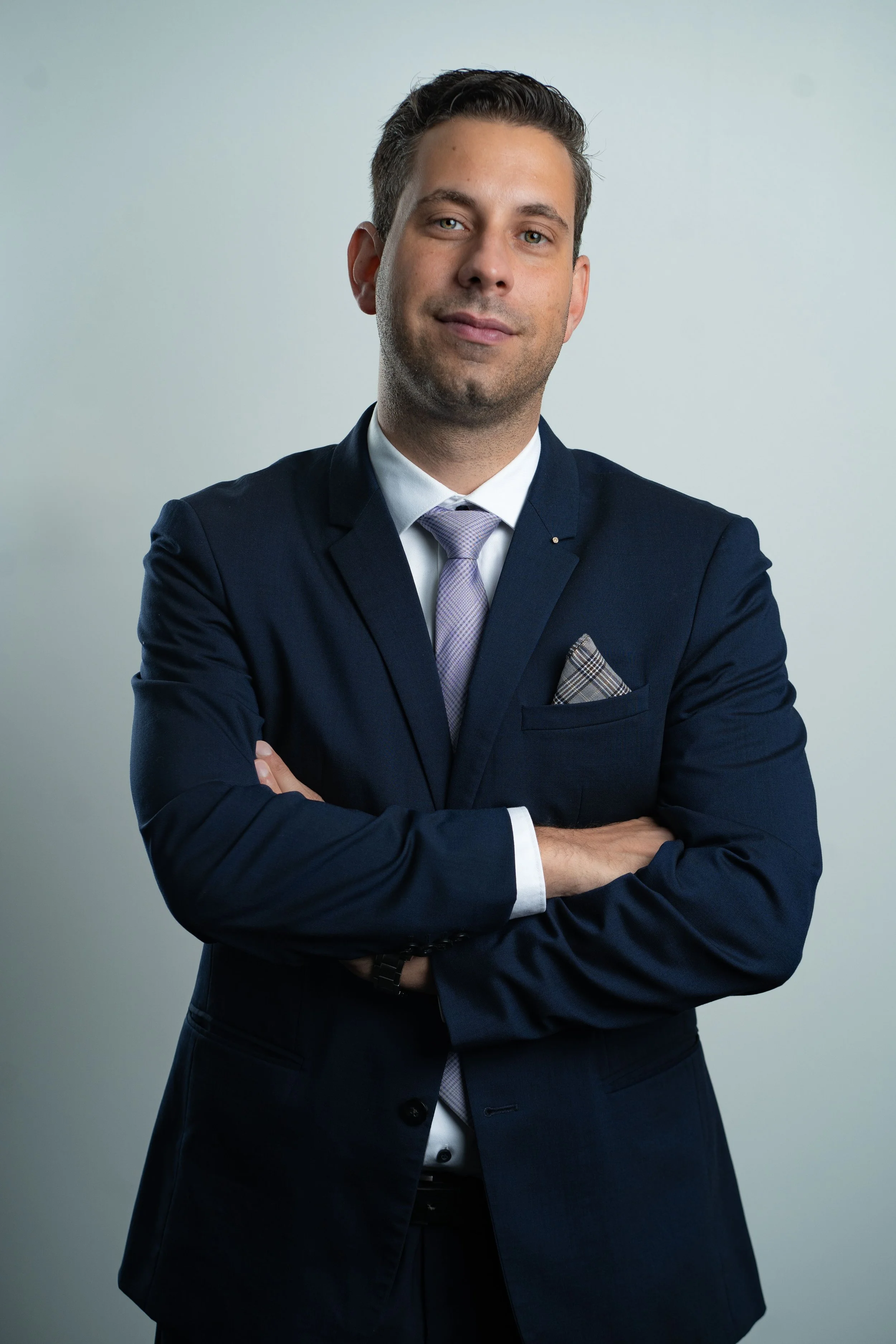 Alexander Jungbluth in dunklem Anzug mit Krawatte und Jackett posiert mit verschränkten Armen vor hellem Hintergrund.