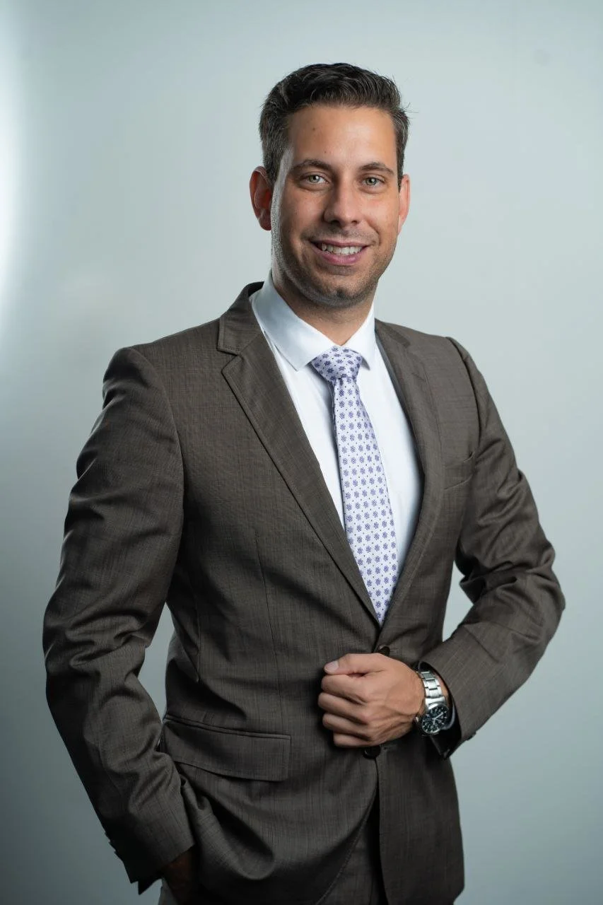 Ein professionell aussehender Mann in Anzug und Krawatte, lächelnd, vor hellem Hintergrund.