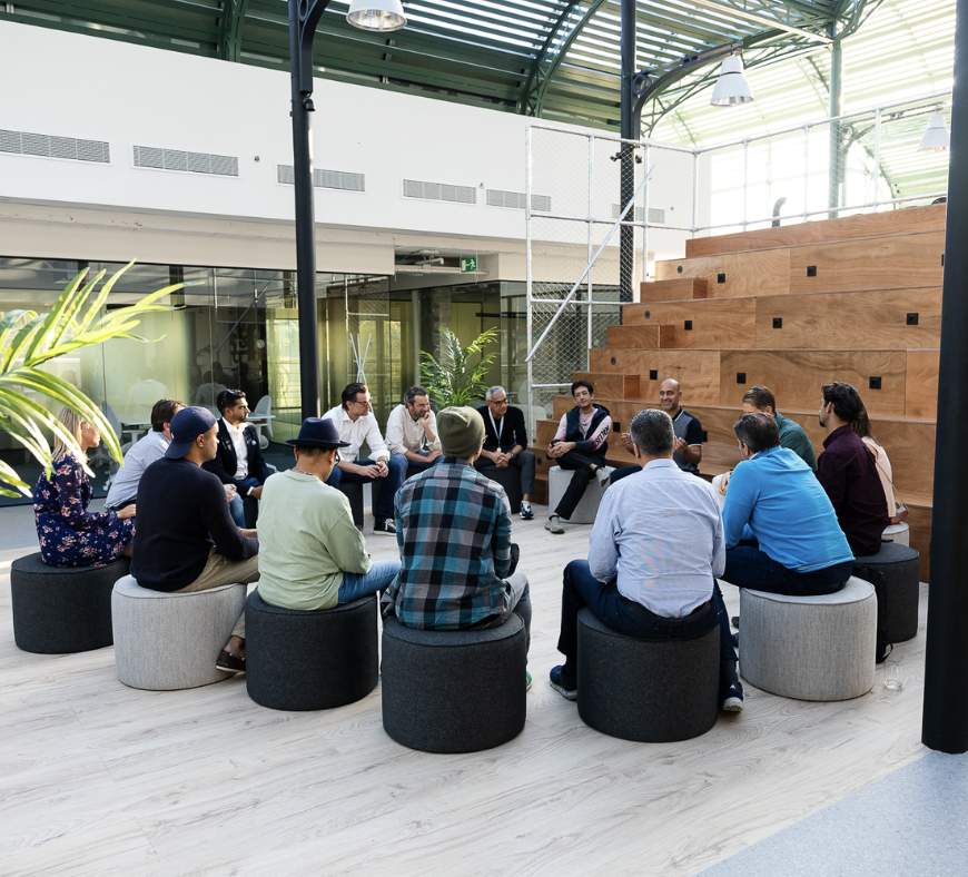 Group of people sitting in a circle on stools in an indoor space with a high glass ceiling, plants, and a staircase.