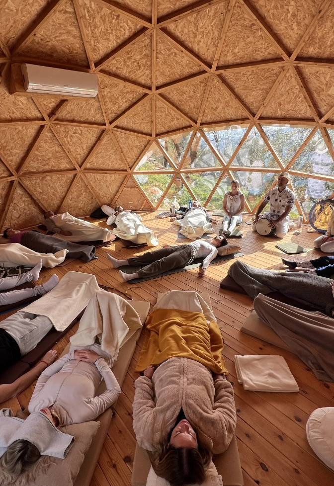 People participating in a guided meditation or relaxation session inside a geodesic dome structure with wooden walls and a large window showing trees outside.