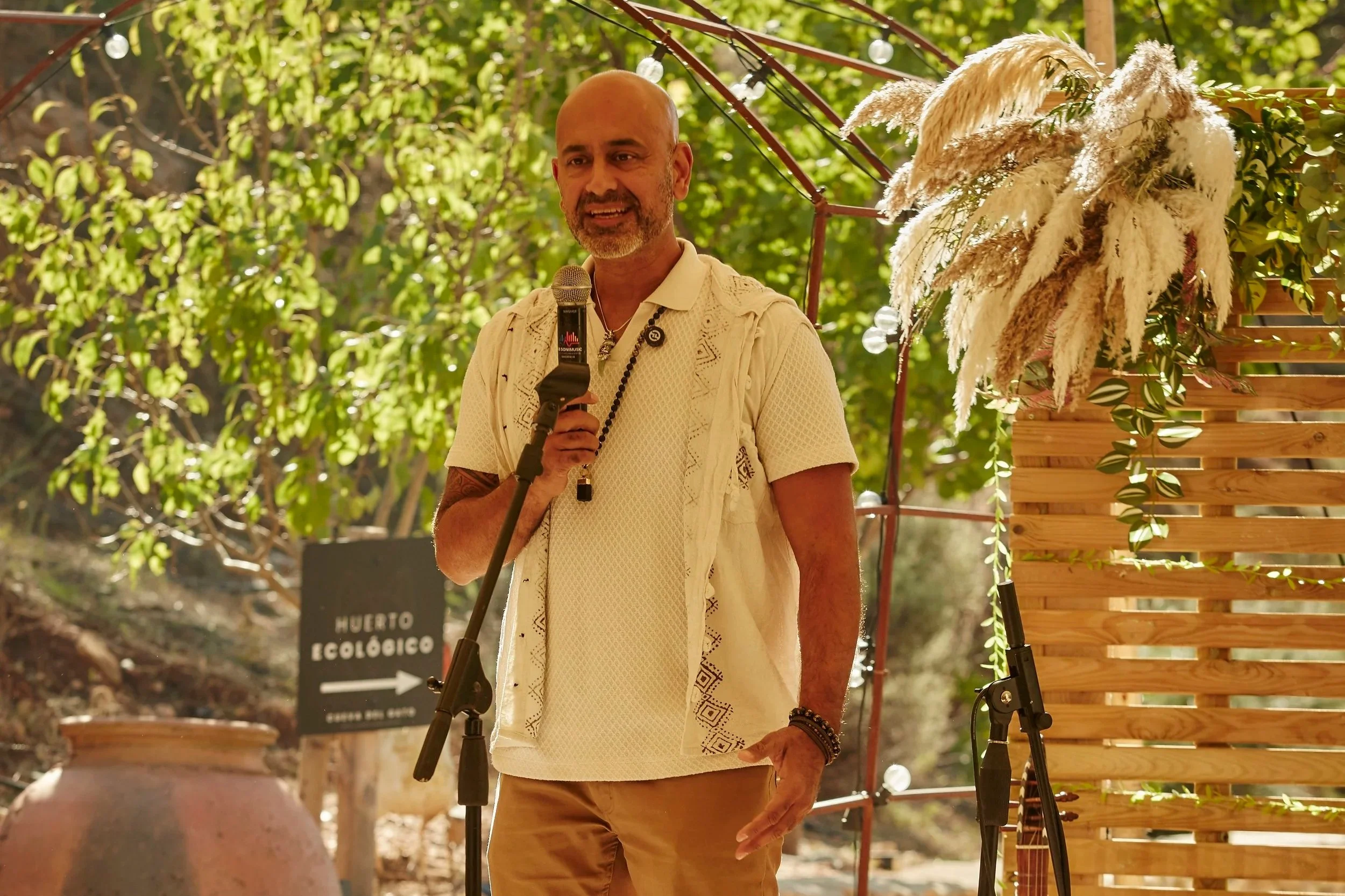 A man holding a microphone speaking outdoors during daytime. He is wearing a cream-colored short-sleeve shirt with lace and embroidery details and tan pants, with bracelets on his wrist. Behind him is a wooden lattice with white pampas grass and greenery. There is a sign pointing to an eco-friendly farm in the background.