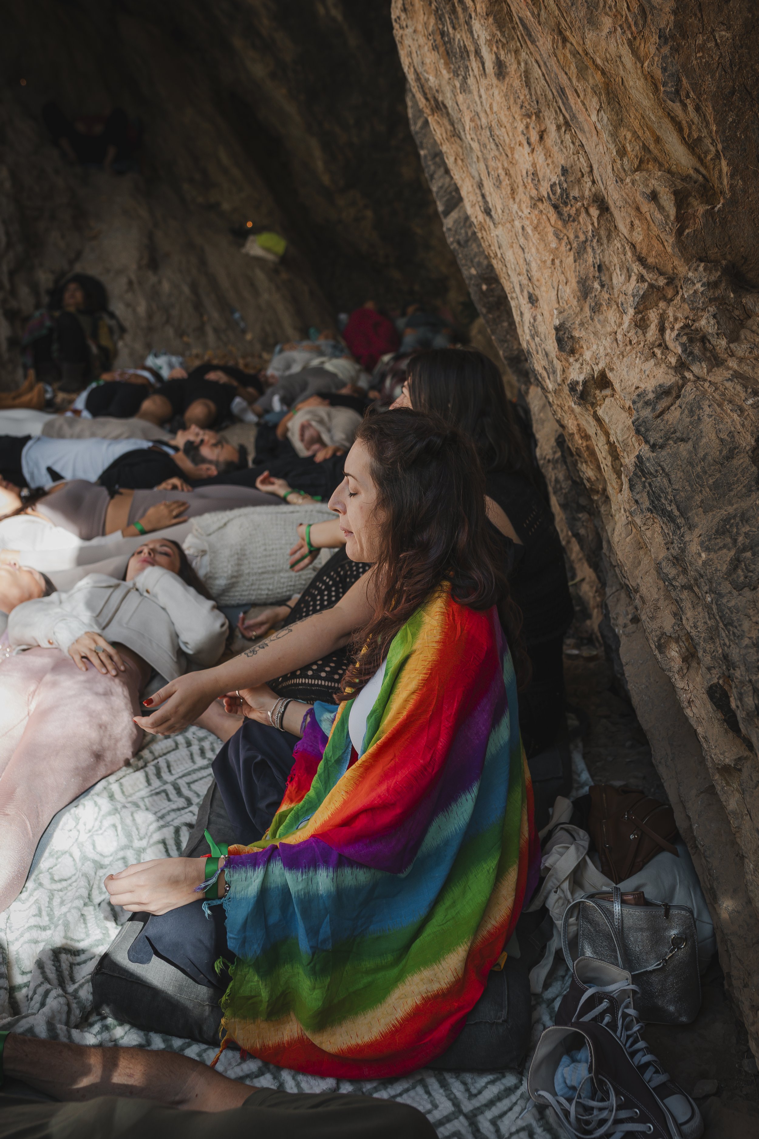 People sitting and lying down in a cave, some with eyes closed, with a woman in rainbow-colored shawl in foreground.