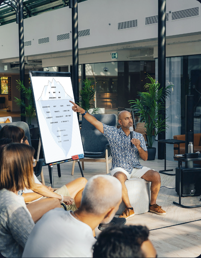 A man in a patterned shirt and shorts sitting on a stool, holding a microphone, and pointing at a presentation board with his right hand while speaking to an audience in a modern, well-lit indoor space with plants and glass walls.
