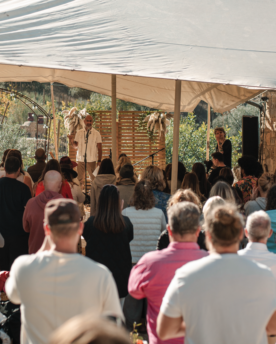 A man speaking into a microphone on a stage decorated with pampas grass and greenery during an outdoor event under a tent.