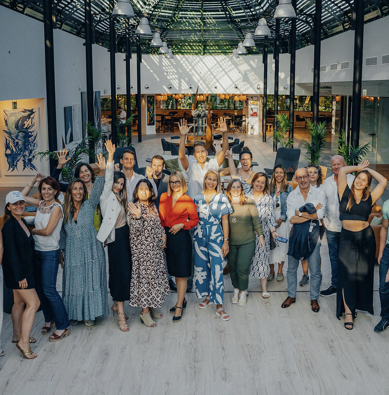 A large group of diverse people smiling and posing together in a spacious, modern indoor setting with artwork and plants.