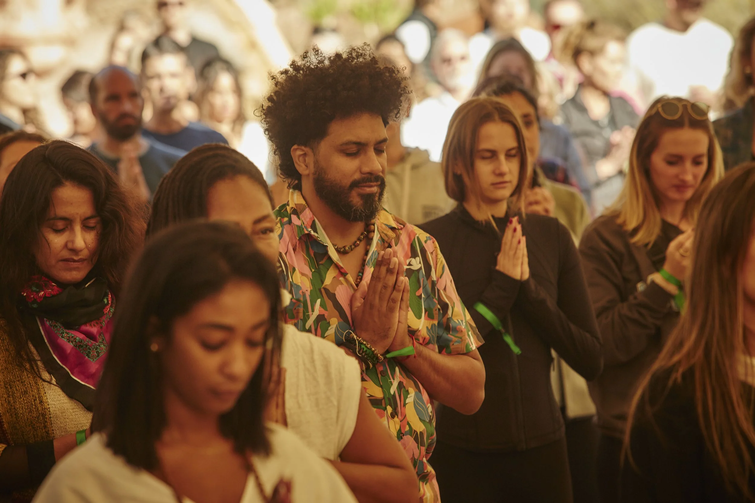 A diverse group of people with eyes closed and hands in prayer or meditation, gathered outdoors in a peaceful setting.