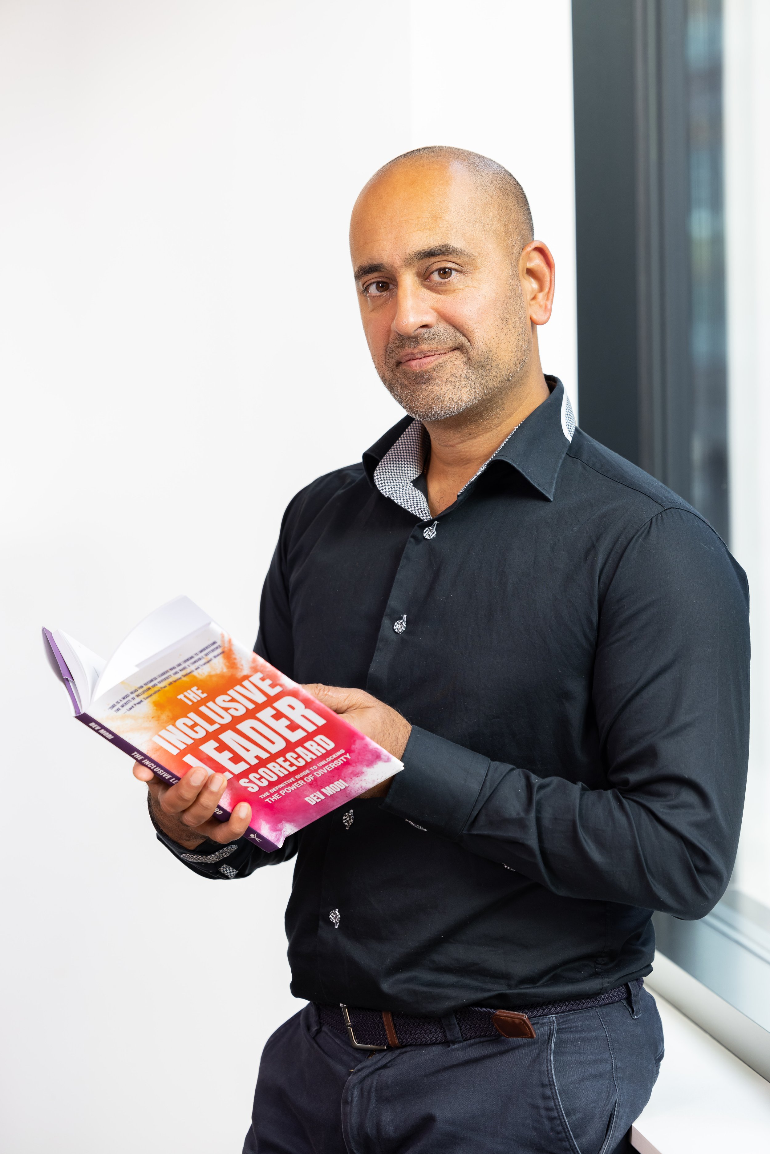 A man with a bald head and gray beard stands near a window, holding a book titled 'The Inclusive Leader Scorecard' and smiling softly.