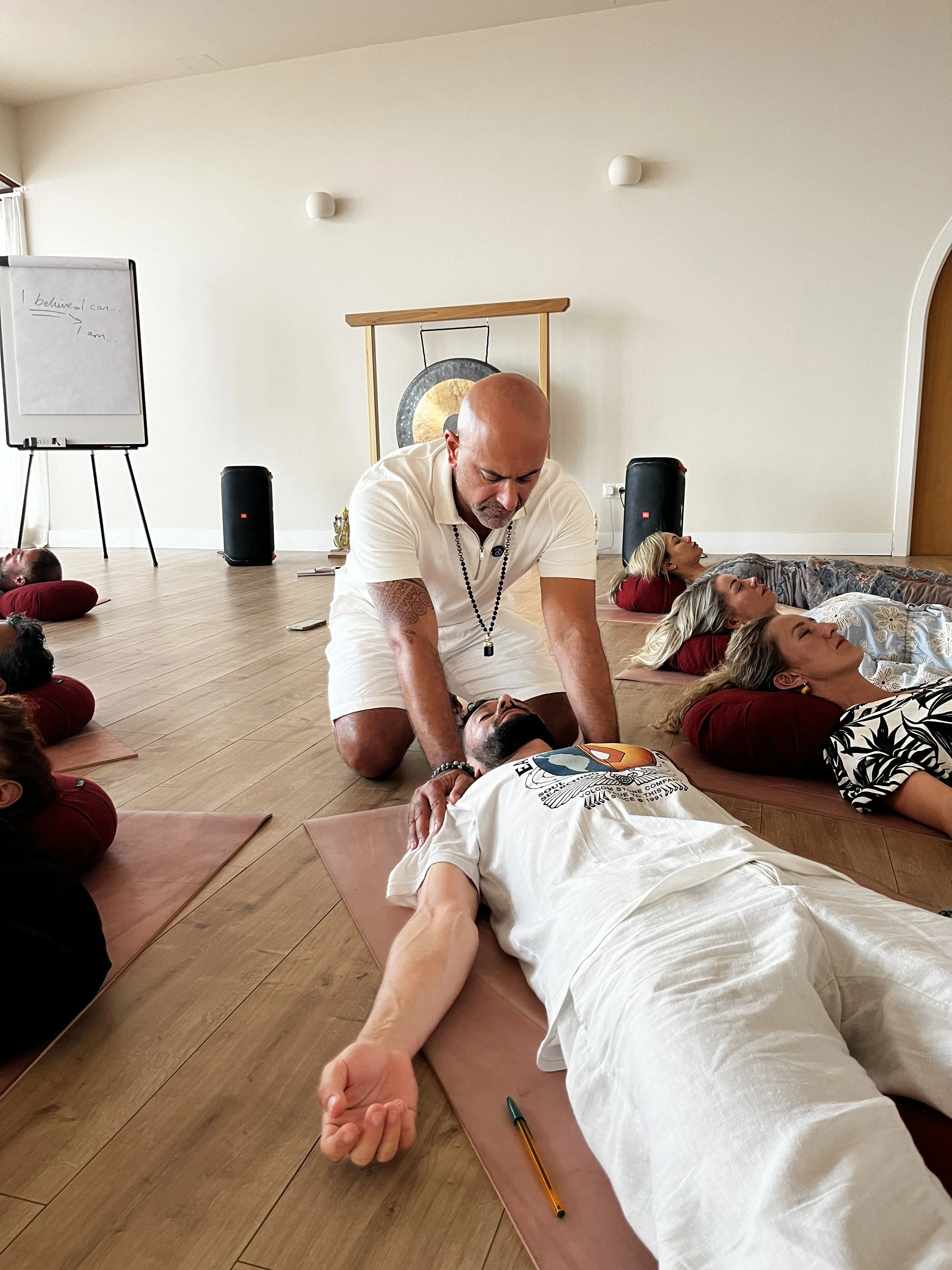 People participating in a group meditation session, lying on mats on the wooden floor with their eyes closed, as a man in a white shirt provides a hands-on healing or energy work.