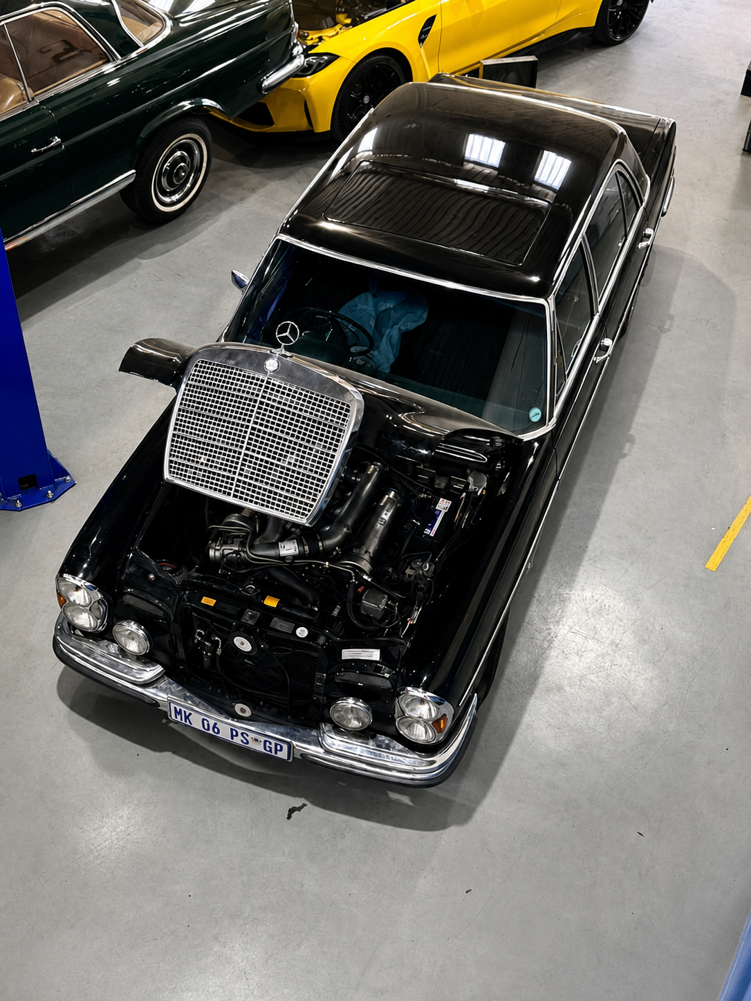 Black vintage Mercedes-Benz car with the hood open showing engine in a showroom or garage, surrounded by other classic and modern cars.