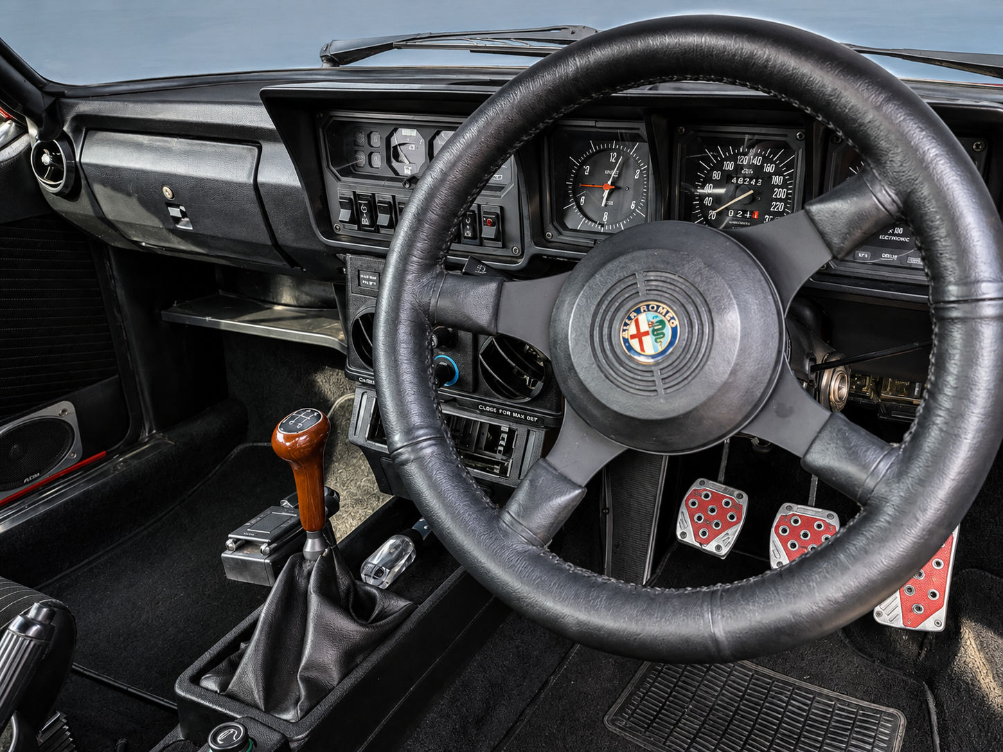 Interior view of a vintage Alfa Romeo car with a black dashboard, manual gear shift with a wooden handle, and sporty red and silver pedals.