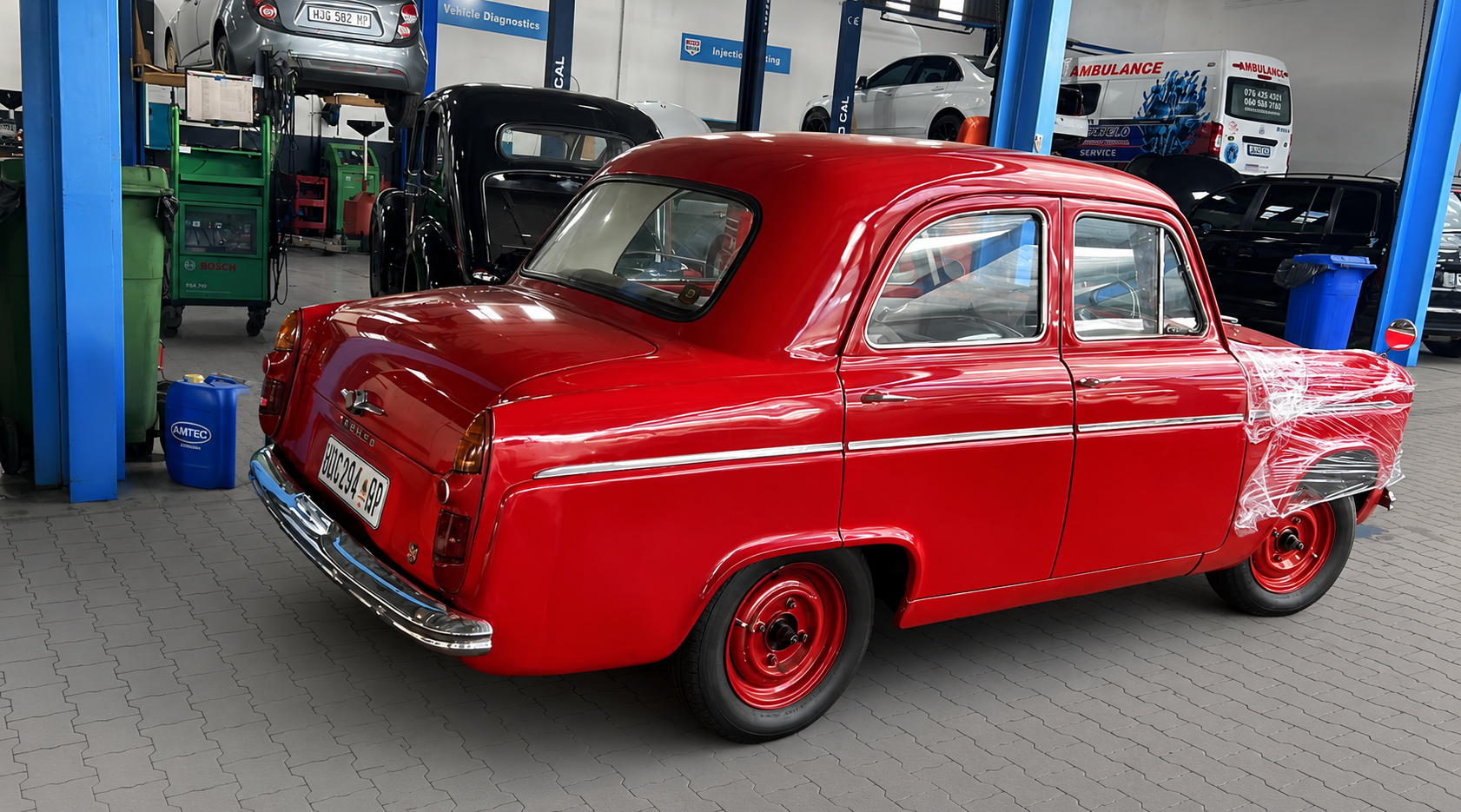 A vintage red car with protective plastic wrapping on the front right fender, inside an auto repair shop or garage.