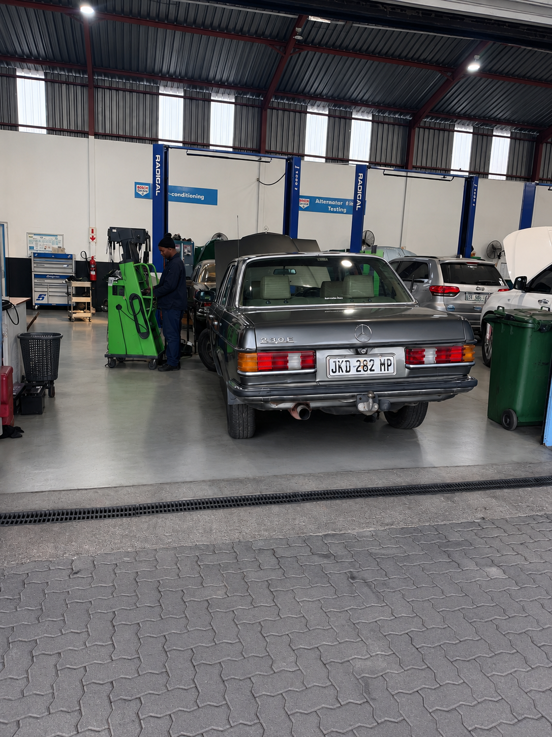 A mechanic working with tools next to a vintage black Mercedes-Benz 230E inside an automotive repair shop, with other cars and equipment visible in the background.