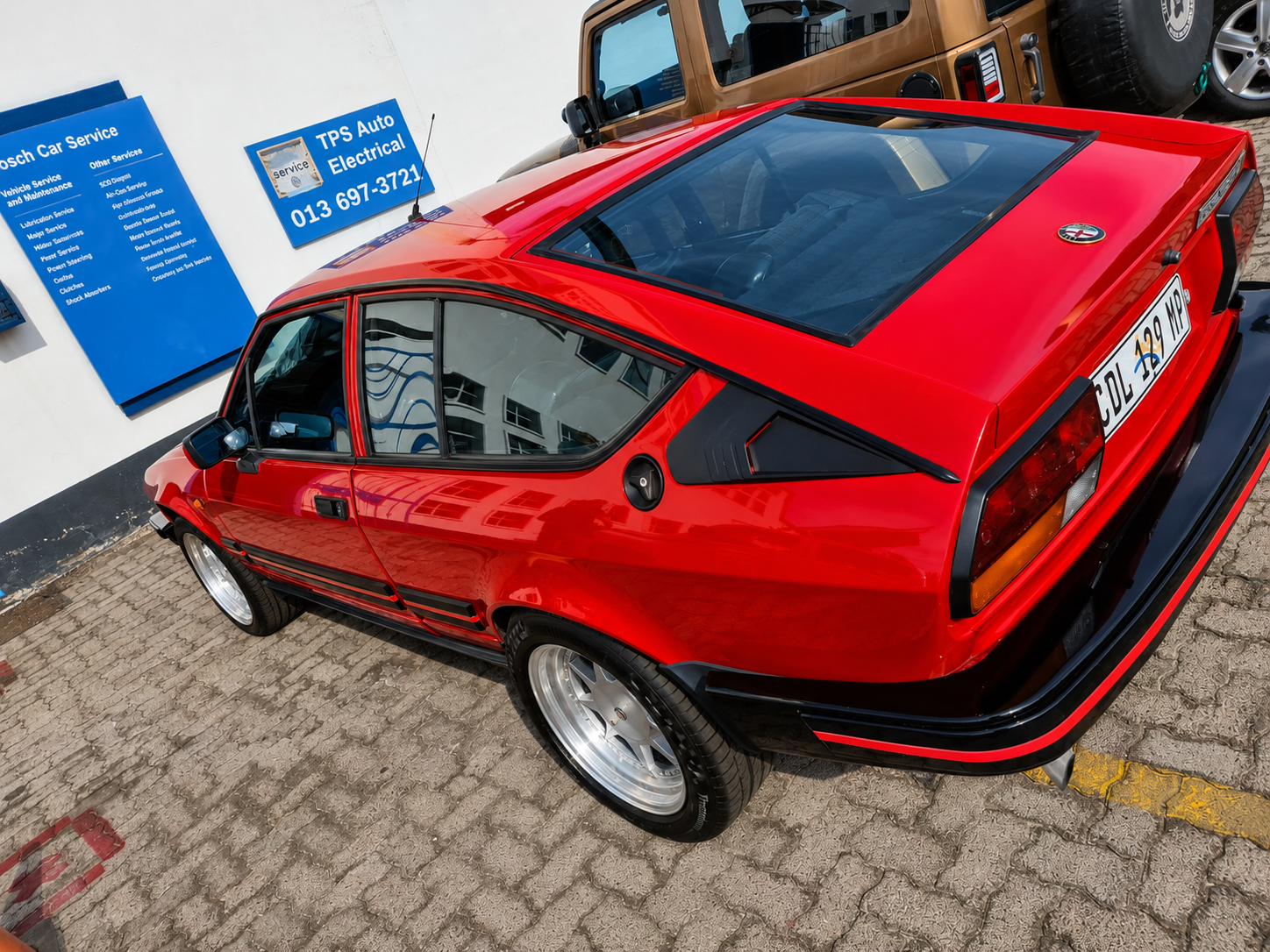 A red vintage car parked on a cobblestone pavement outside a building with a blue sign and white wall. The car has a black trim and a large rear glass window.