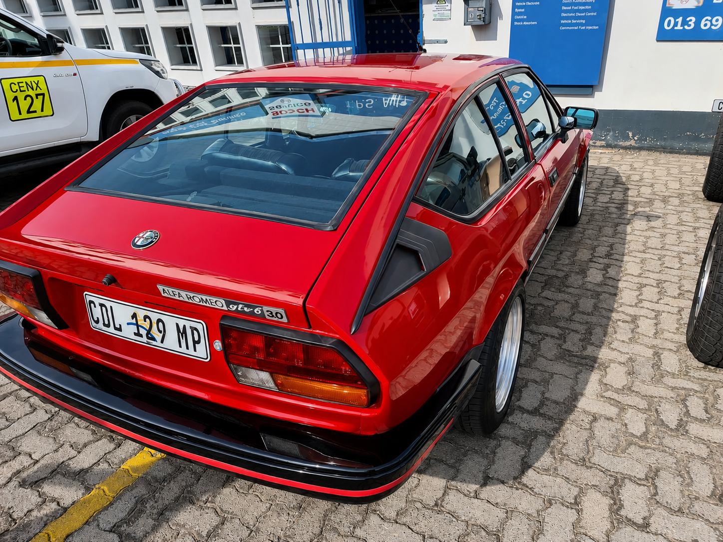 Red vintage Alfa Romeo GTV 6 parked on cobblestone pavement, with a white vehicle and a building in the background.