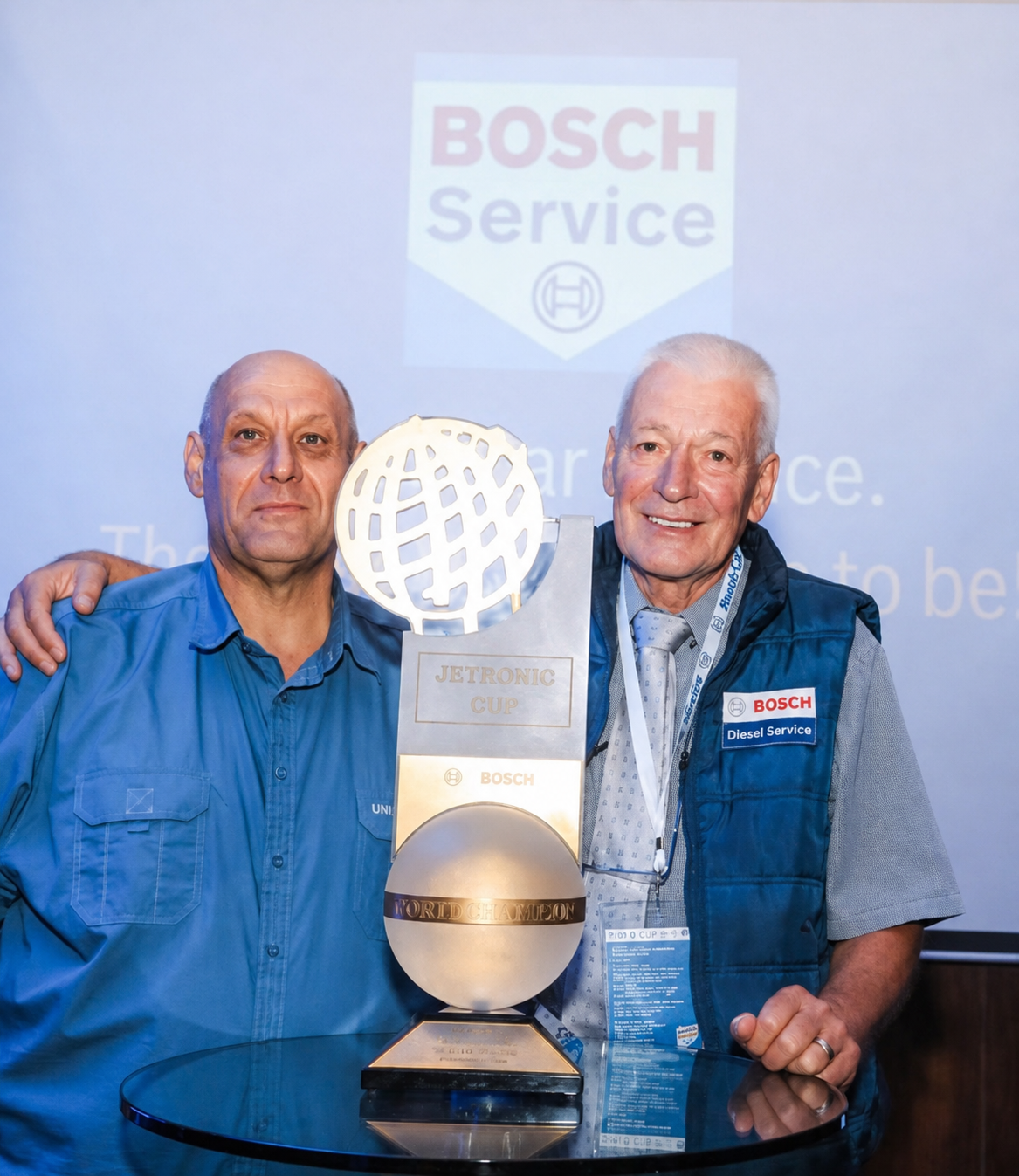 Two men standing in front of a Bosche Service sign, holding a Jetronic Cup trophy with a globe on top. The man on the left is wearing a blue work shirt, and the man on the right is wearing a blue vest with a Bosch Diesel Service logo. They are smilin