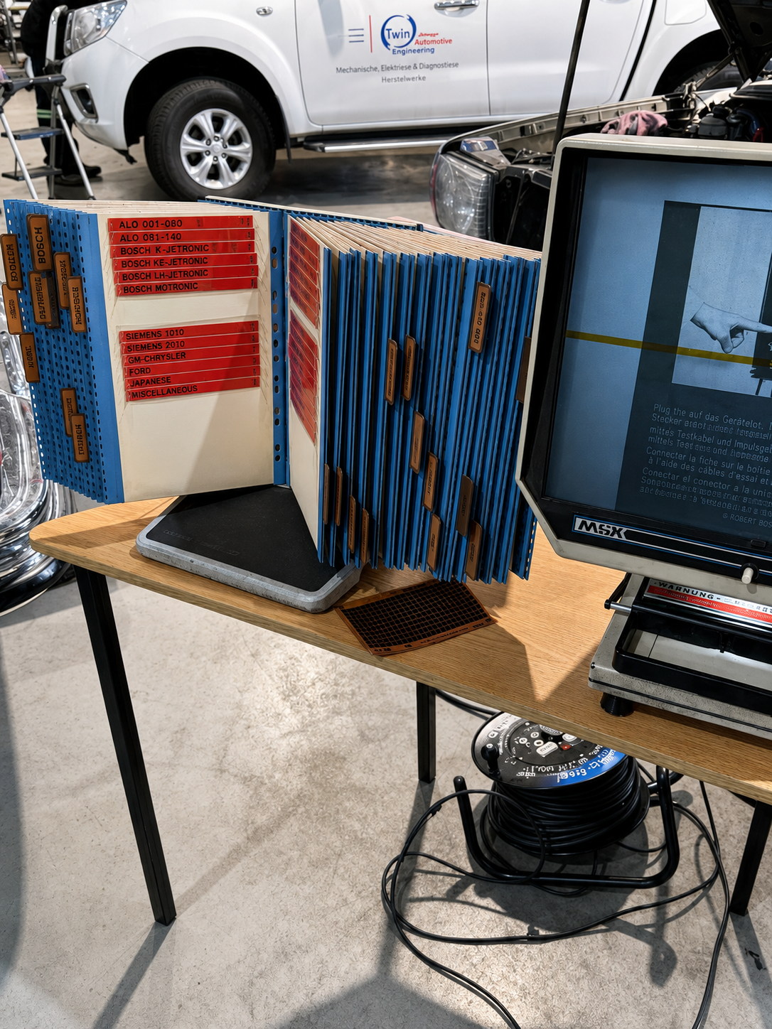 A workstation with a computer monitor displaying testing instructions, a pegboard with labeled automotive parts and test kits, a computer scale, and a reel of extension cords on a wooden table, with cars in a garage or workshop in the background.