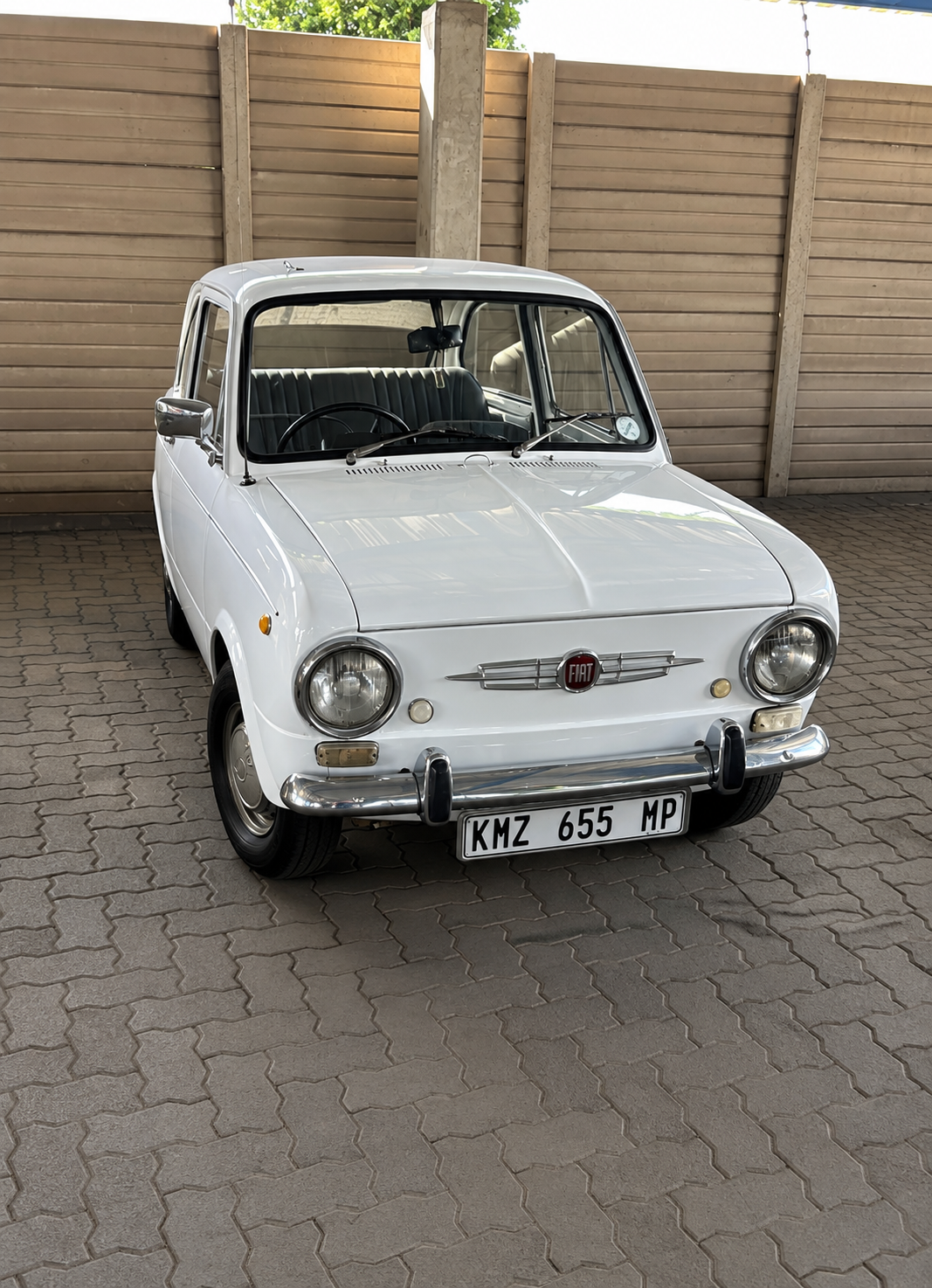 A vintage white Fiat 500 car parked on a paved surface with a wooden fence and greenery in the background.