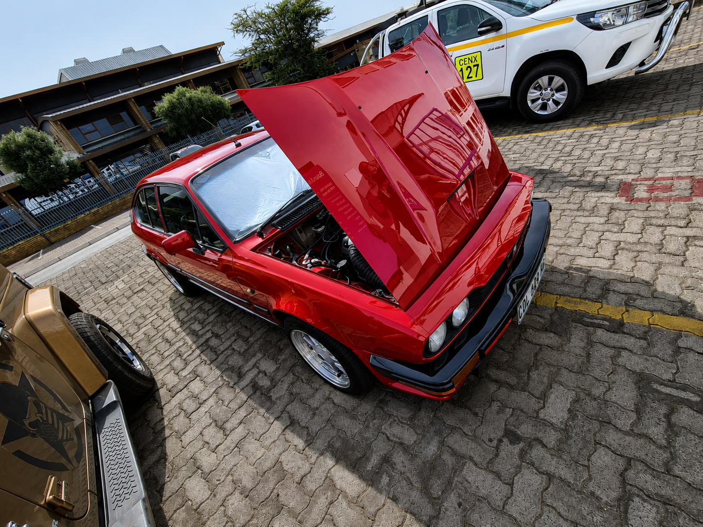Red classic car with its hood open, parked in a lot. There are other cars around, including a white vehicle with a yellow license plate and a beige vehicle with a black decal.