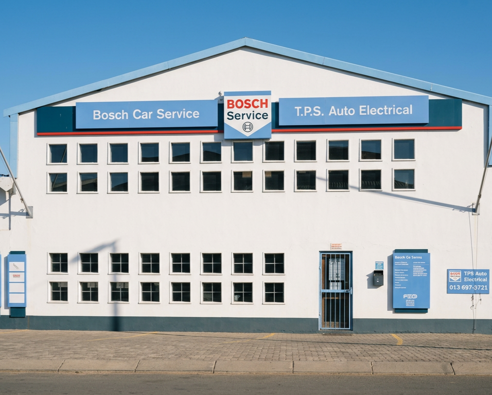 Front of a white building with blue signs for Bosch Car Service and T.P.S. Auto Electrical, featuring a small barred door and several windows under a clear blue sky.