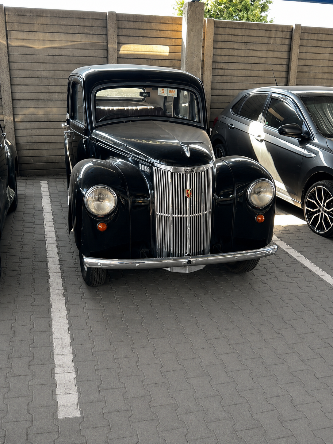 A black vintage car parked in a parking lot between two modern cars, with a wooden fence in the background.