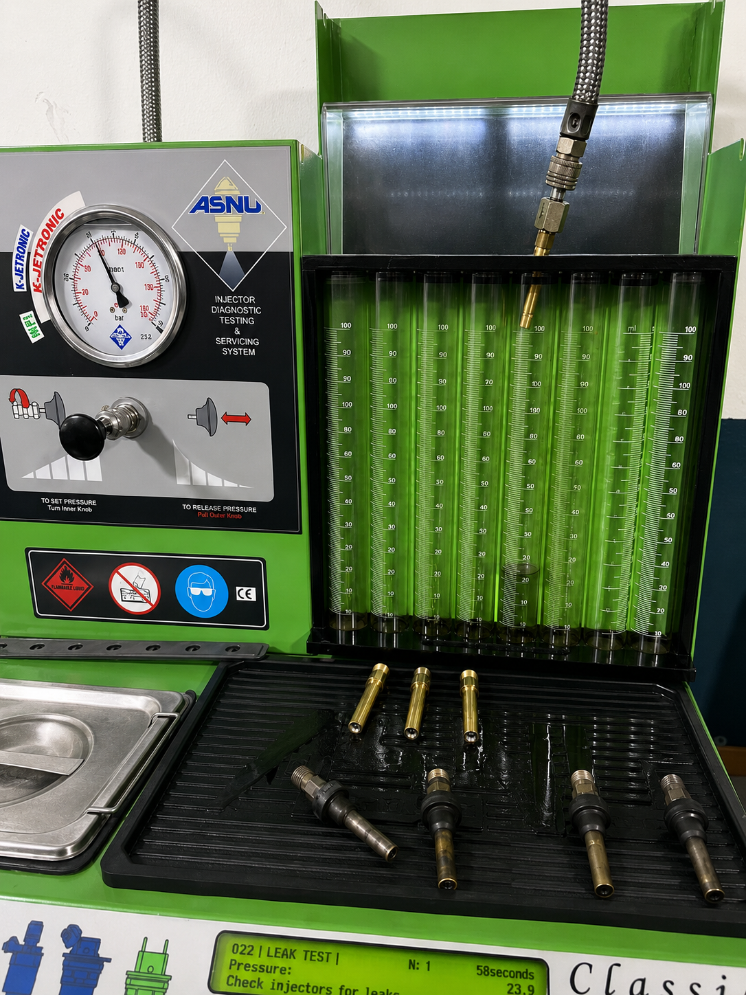 A fuel injector testing and cleaning machine with a pressure gauge, several transparent cylinders, and fuel injector components on a black tray.