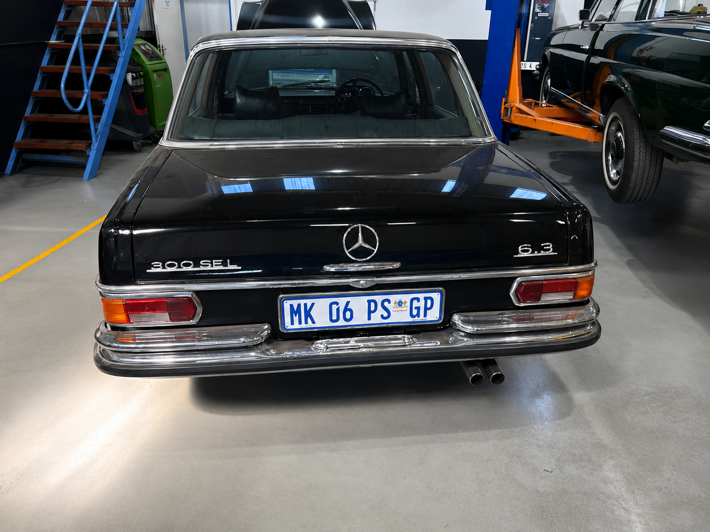 Black vintage Mercedes-Benz 300 SEL 6.3 parked indoors with hospital parking plate. Surroundings include a green recycling bin, a blue staircase, and a green vehicle on a lift.