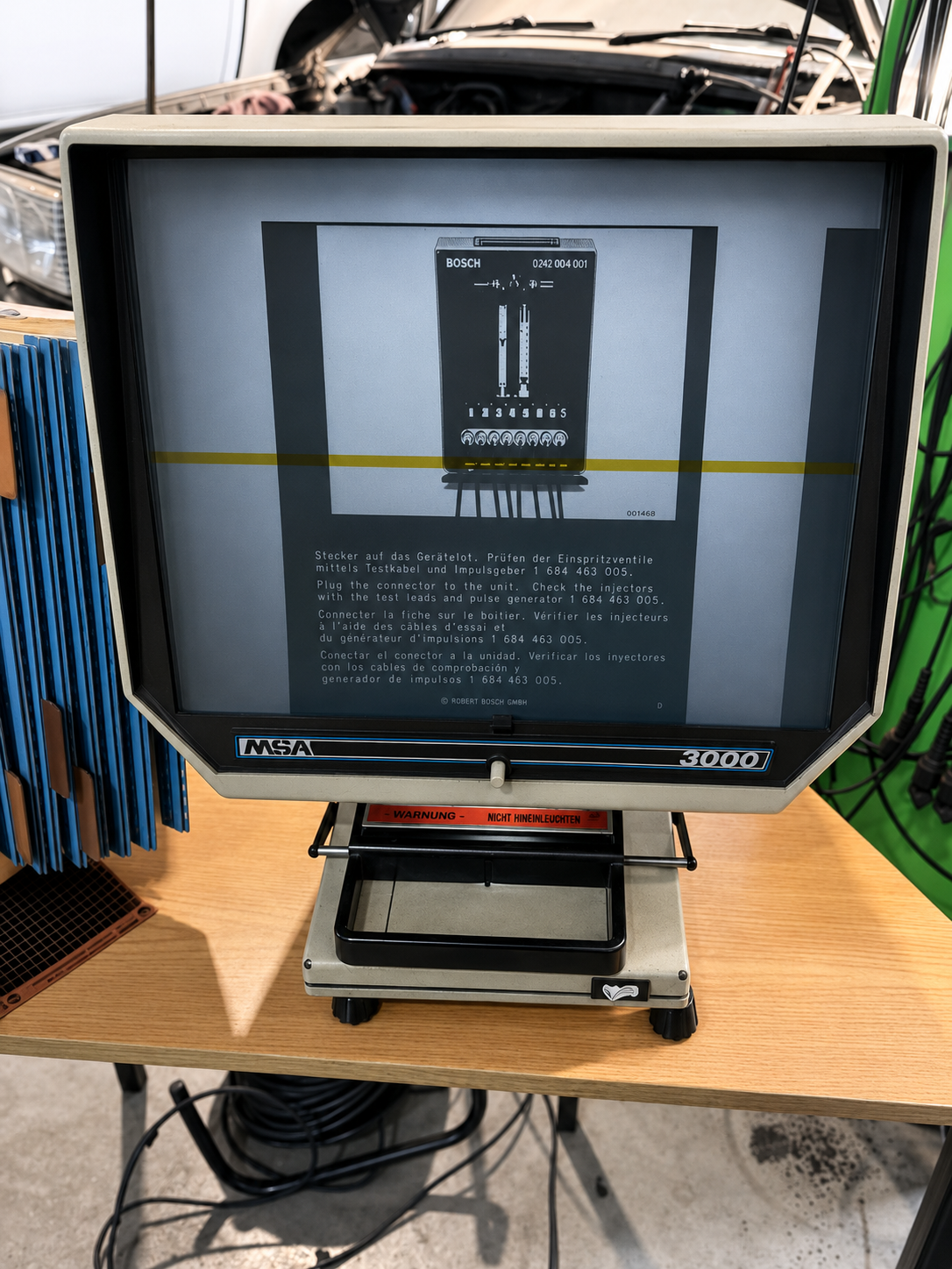 Display screen showing instructions for checking fuel injectors with a test lead and pulse generator, with surrounding tools and vehicle in the background.