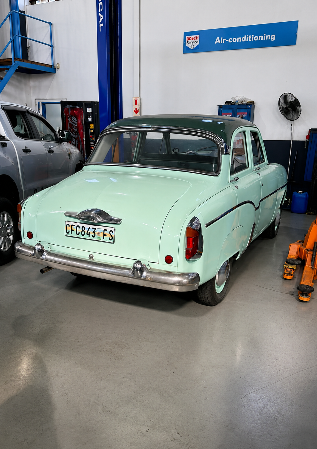 A vintage pale green car with a dark roof inside an auto repair shop, next to a silver vehicle and various auto equipment.