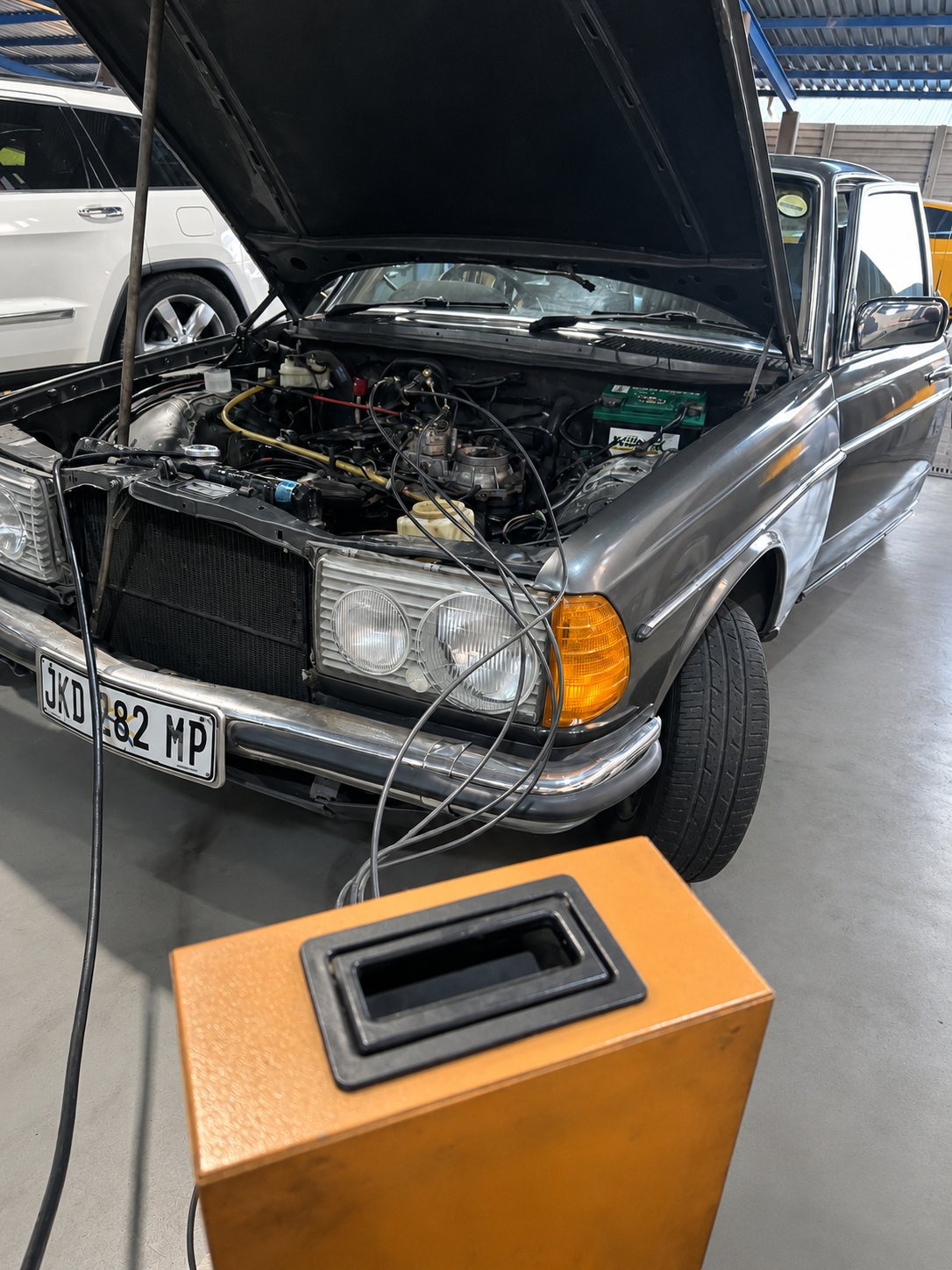 A vintage car with its hood open in a garage, connected to testing equipment on a stand.