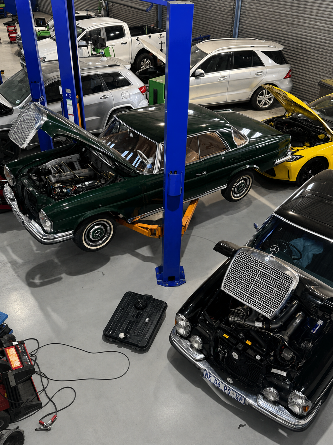 A workshop with several cars, including a classic green car on a lift with its hood open, a yellow car with its hood open, and various other vehicles parked around. There are tools and equipment on the floor.